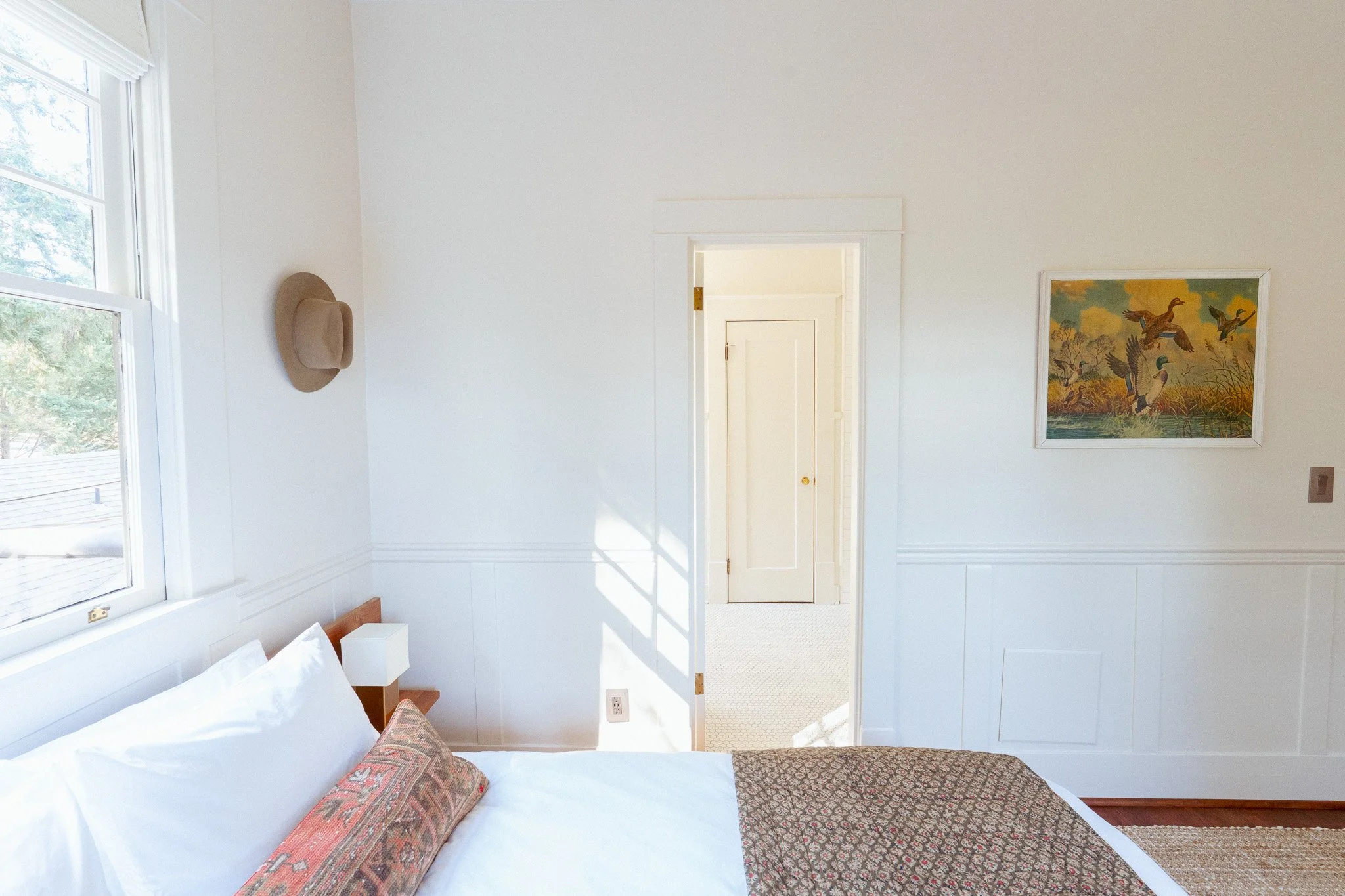 A bedroom with white walls and a window on the left, a bed with white pillows, a decorative pillow, and a patterned bedspread. There is artwork of ducks on the wall and a beige hat hanging on the wall. A door leads to another room.
