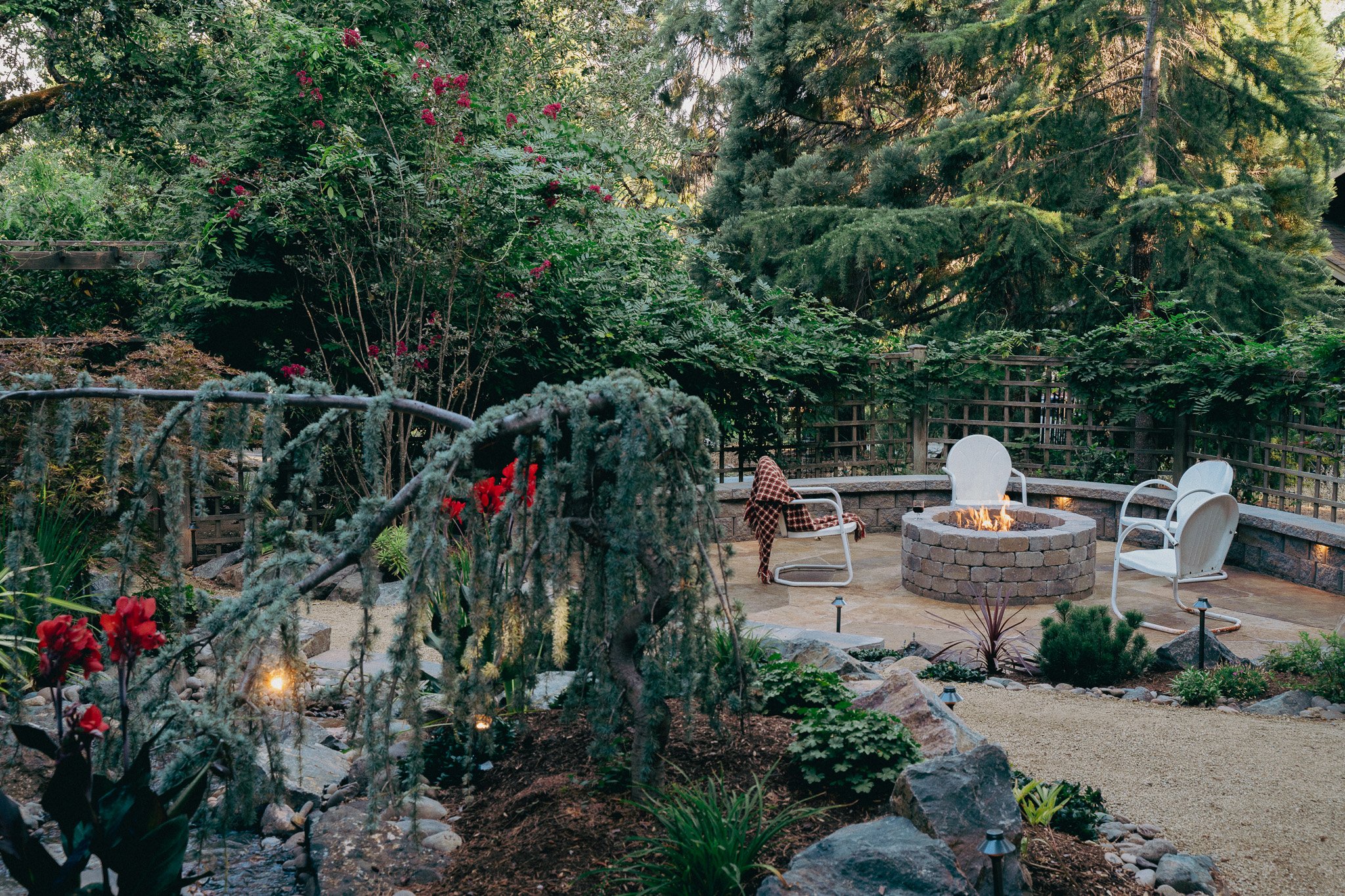 A backyard patio with a fire pit, surrounded by white chairs, with lush green trees and flowering bushes.