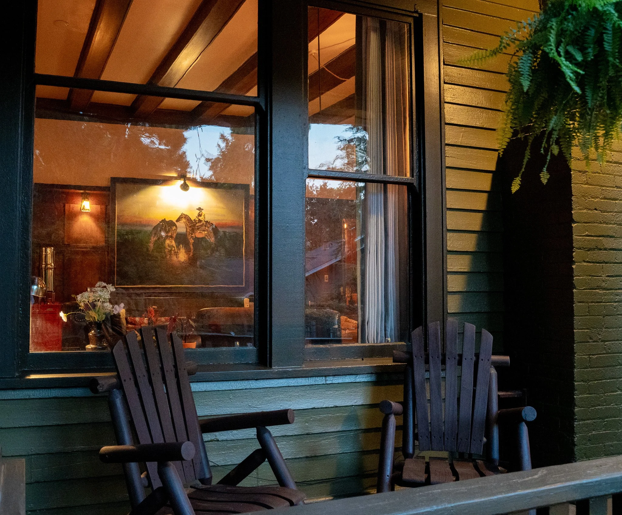 view of a house window with black framing, digital clock on the windowsill, and a painting of horses inside. A green fern in a black pot is mounted on the yellow wooden exterior wall, with outdoor chairs in front.