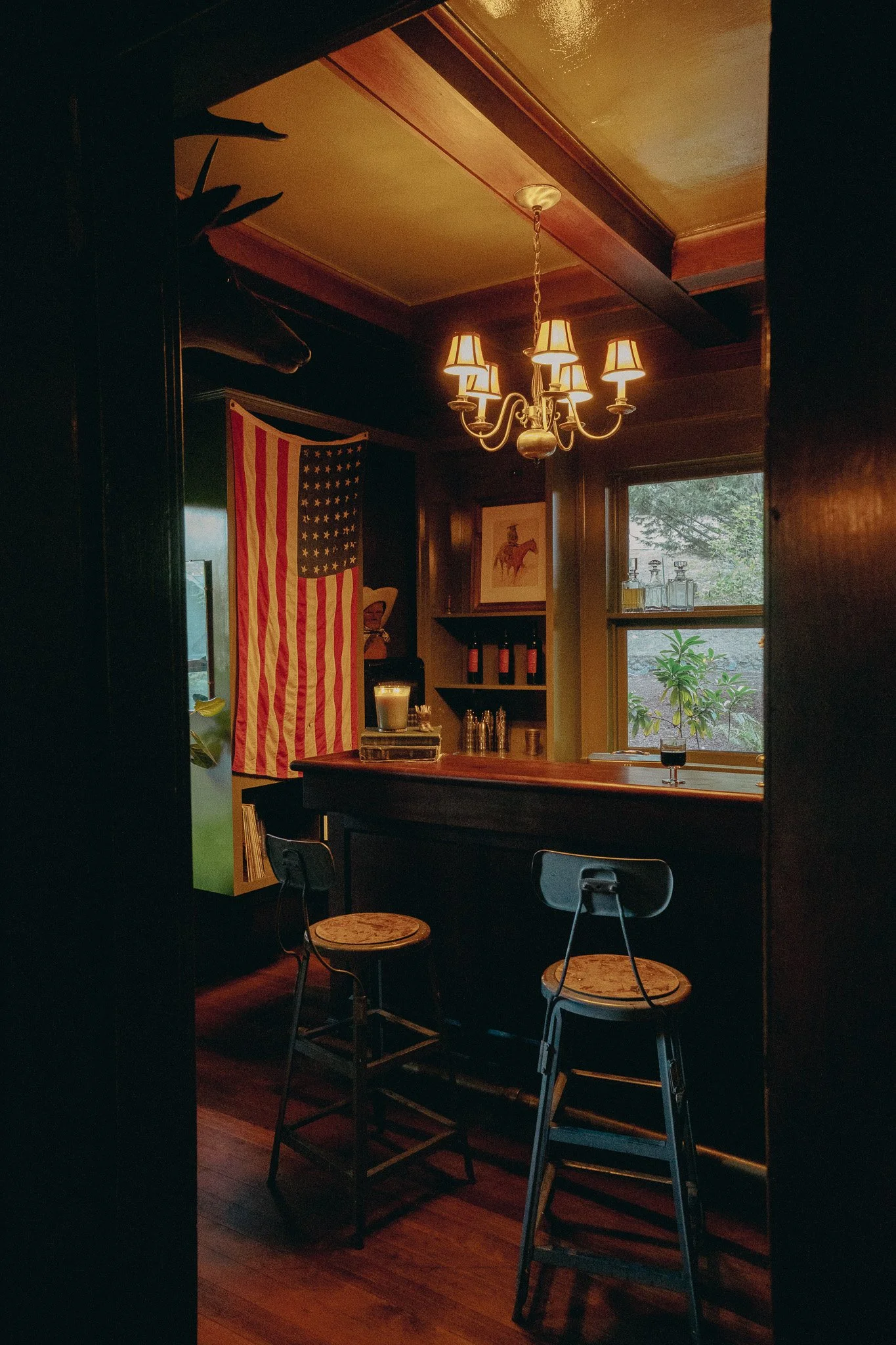 A cozy dining area with wooden accents, featuring a chandelier with five lamps, a window showing greenery outside, and an American flag hanging on the wall.