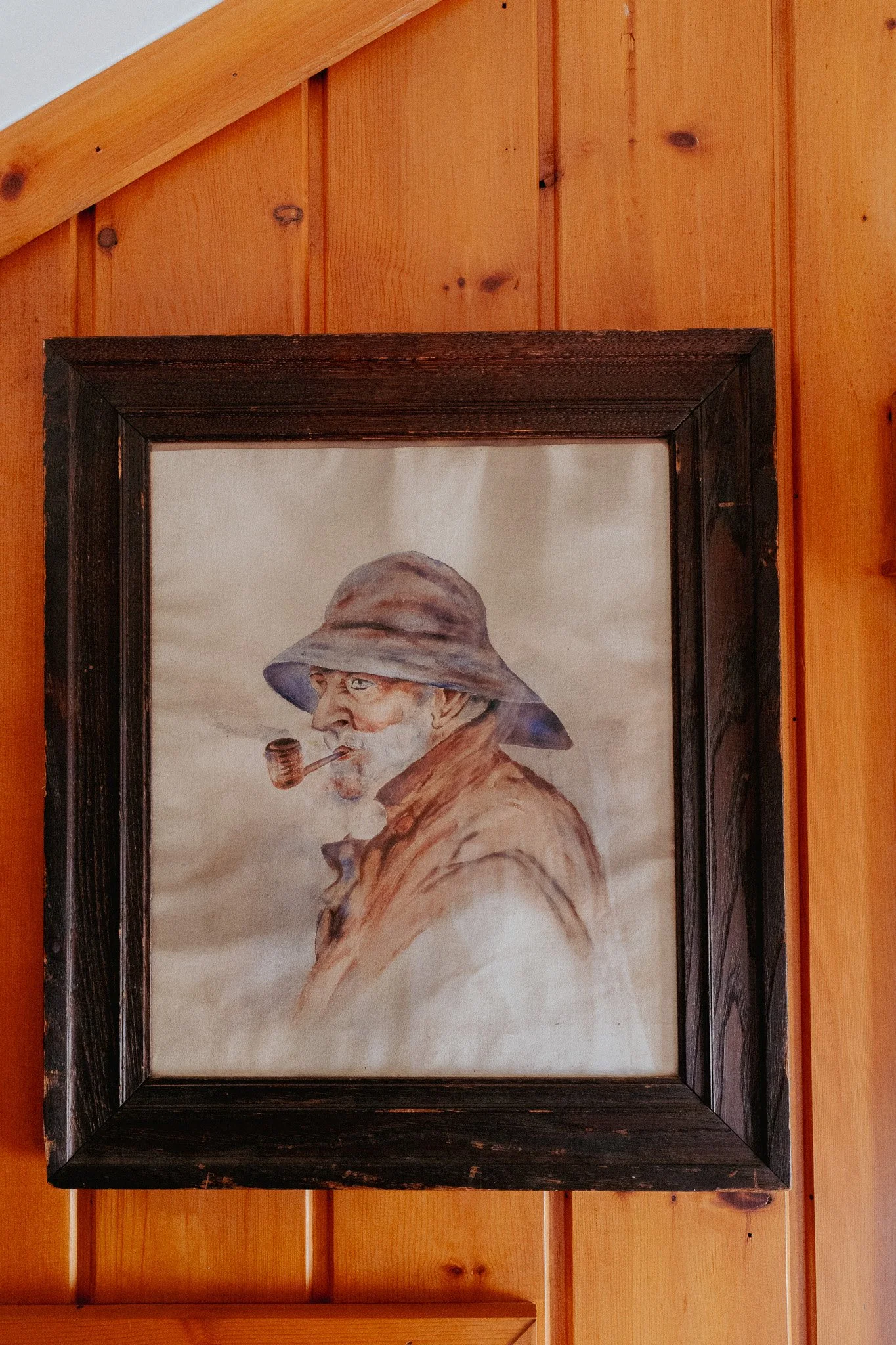 Painting of an elderly man with a white beard smoking a pipe, wearing a wide-brimmed hat and a scarf, framed and hung on a wood-paneled wall.