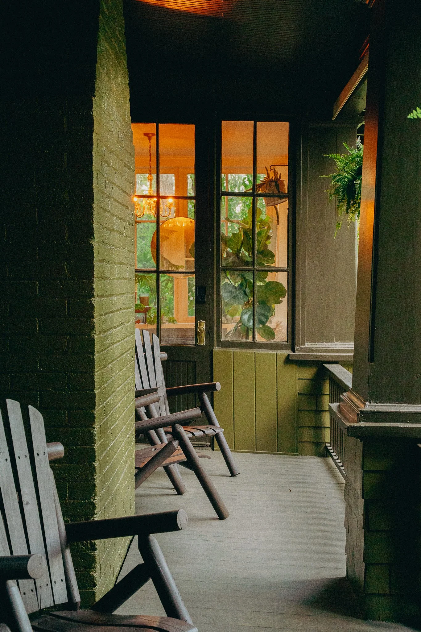 A porch with a row of wooden chairs facing a large window. The window reveals an interior with a chandelier, potted plants, and sheer curtains. The porch has dark painted walls and a light-colored wooden floor.