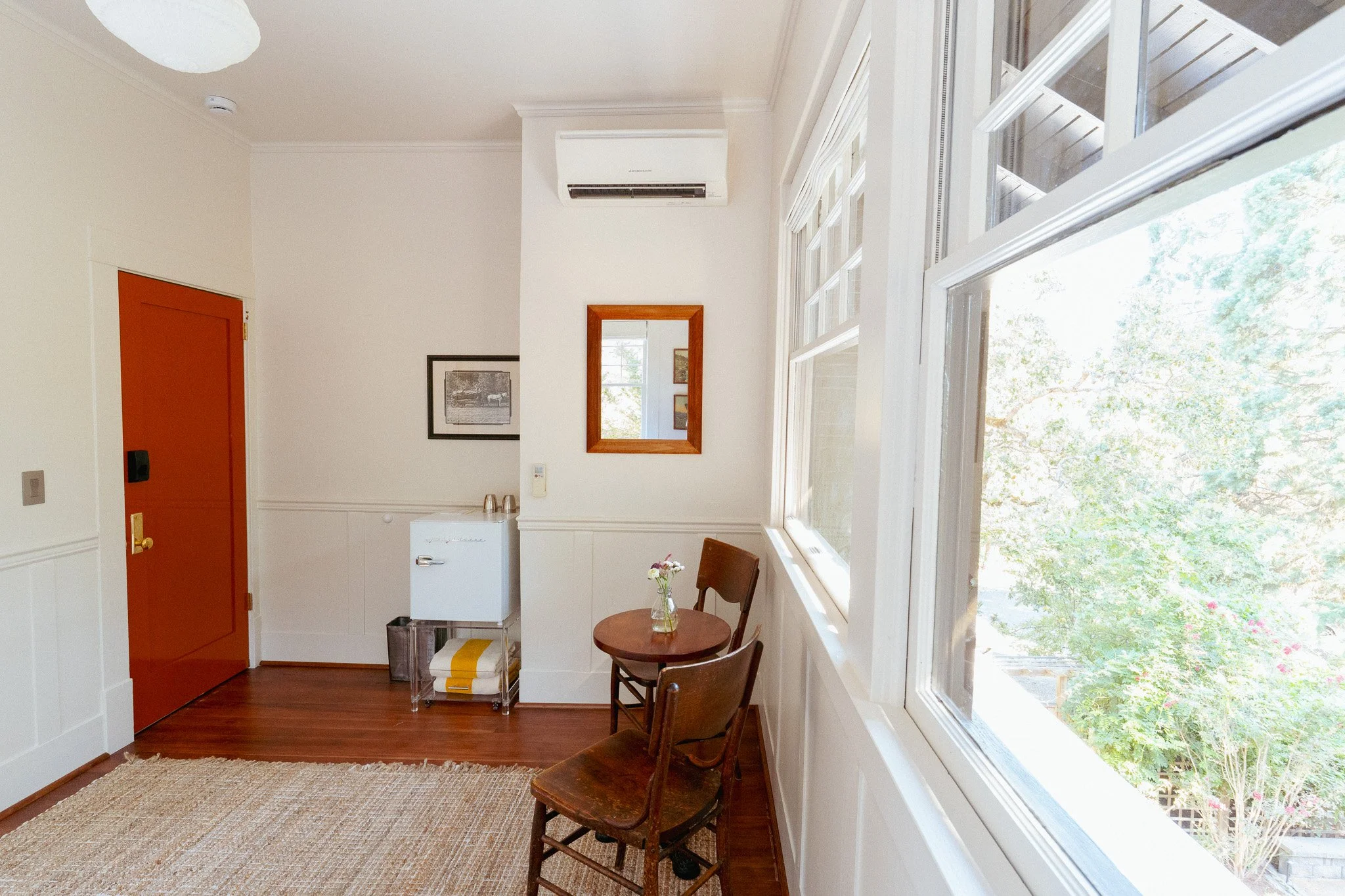 Bright room with large windows showing trees outside, white walls, hardwood floors, an orange door, two wooden chairs, small white refrigerator, wall-mounted air conditioner, framed artworks, and a mirror.