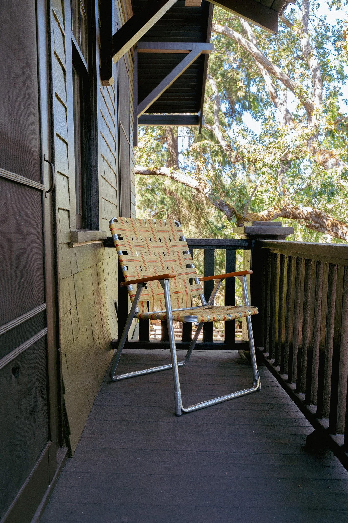 A balcony with two striped outdoor chairs facing each other, surrounded by wooden railings and wall, with trees and blue sky visible in the background.