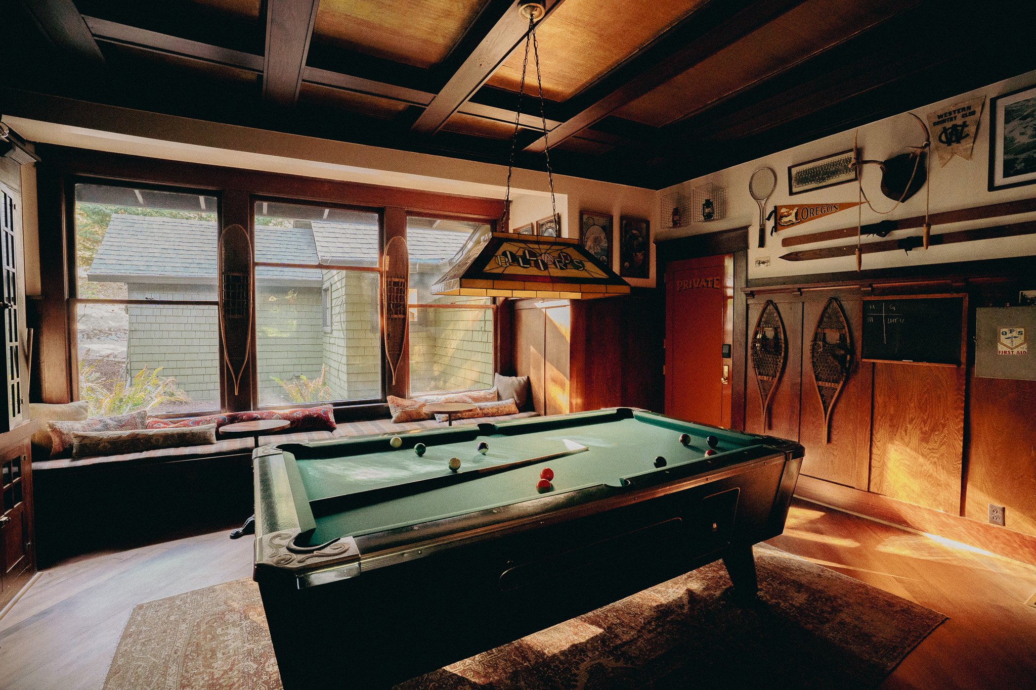 Interior of a room with a pool table, large windows, and wall decorations including skis, a tennis racket, and vintage posters, with wooden ceiling and floor.