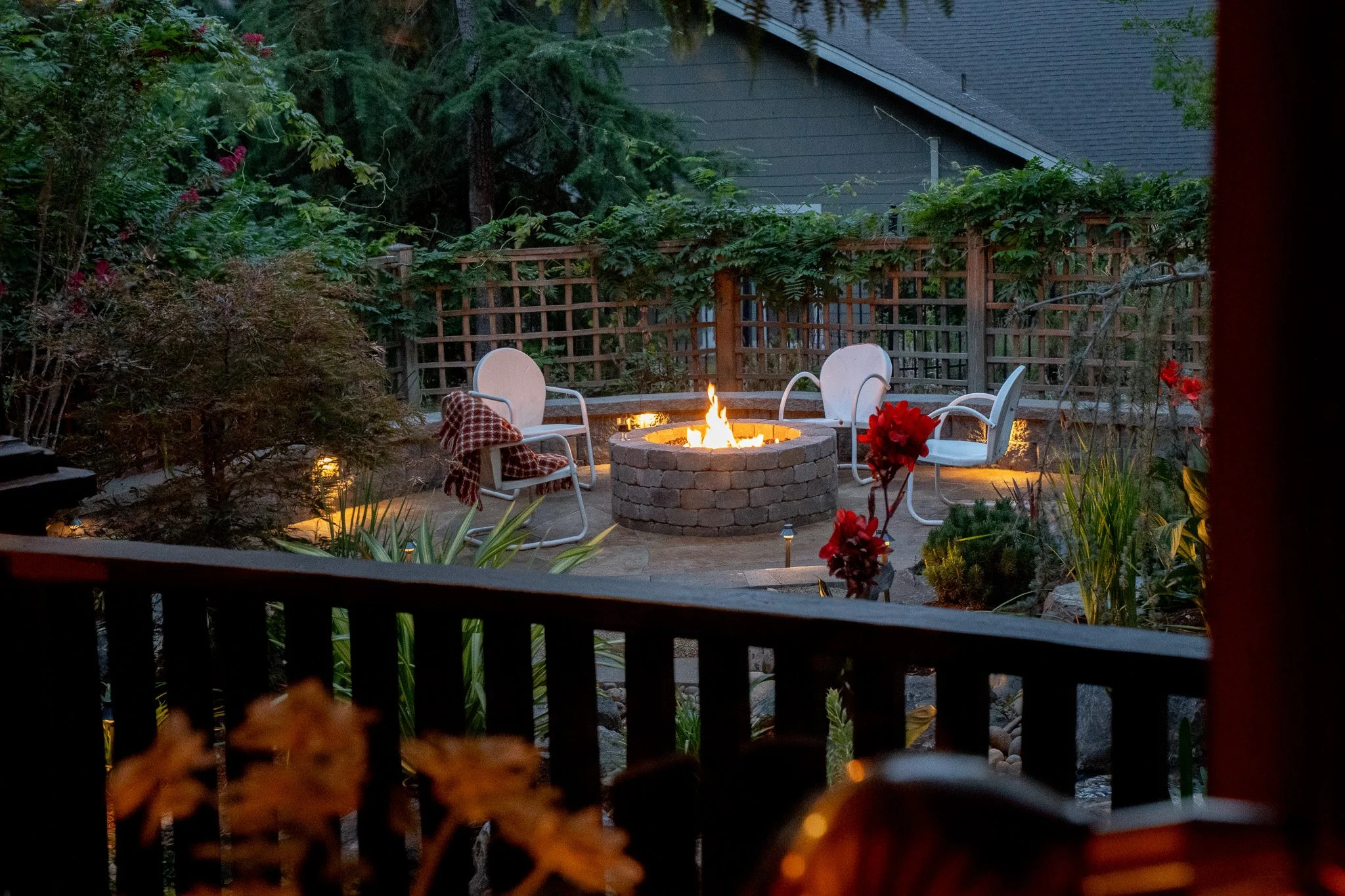A backyard patio with a fire pit, surrounded by four white chairs, and lush greenery including plants and trees. The scene appears to be early evening or dusk.