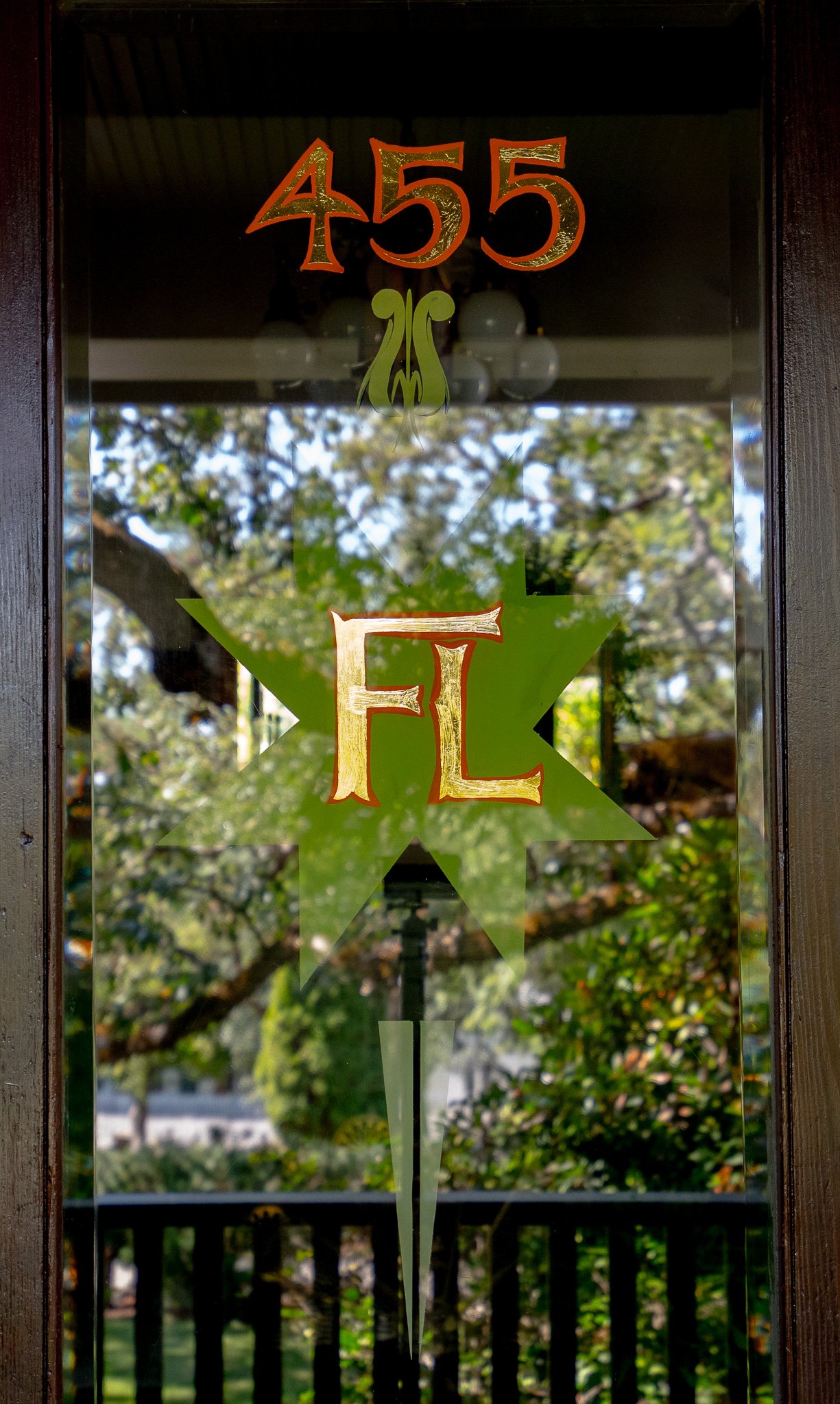 Glass door with house number 455, decorated with a green star and a large letter E. Reflections of trees and a black fence are visible through the glass.