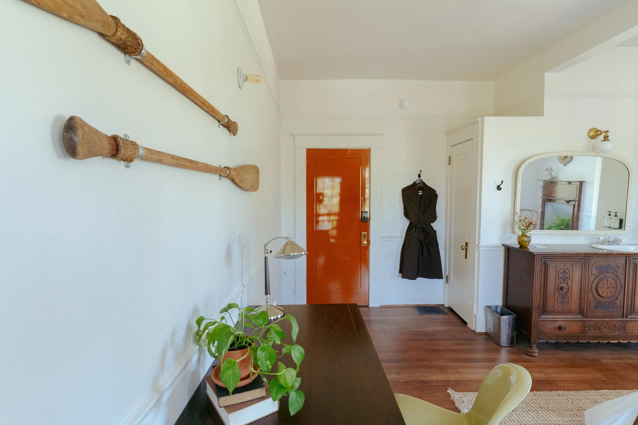 Interior view of a cozy living area featuring a white wall with three old wooden paddles mounted as decor, a red front door, a mirror above a wooden sideboard, a black coat hanging on a hook, a table lamp, a green potted plant, and hardwood flooring.