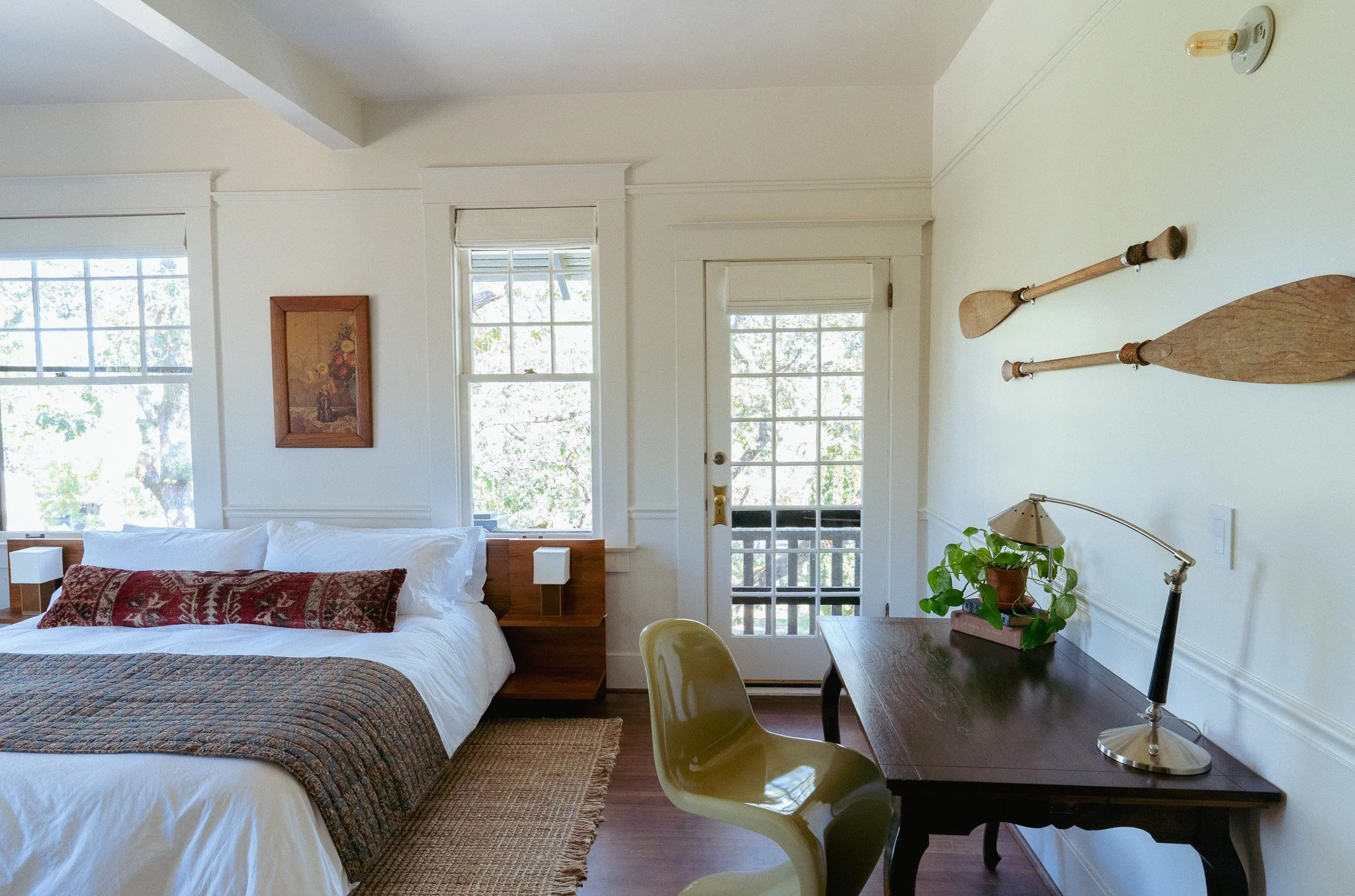 A cozy bedroom with a white bed, patterned quilt, and a pillow with a red and brown design. There is a wooden bedside table with small lamps on each side of the bed. The room has large windows and a glass-paneled door letting in natural light, with trees visible outside. On the wall, there are two wooden oars mounted as decoration and a framed floral painting. There is a dark wooden desk with a green plant in a pot and a silver desk lamp, along with a light green chair.
