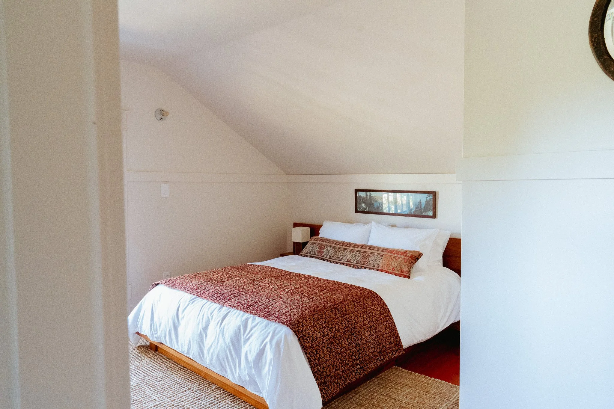 A bedroom with a bed covered in white linens and a patterned bed runner. There is a framed picture hanging on the wall above the bed, and a small nightstand or lamp is partially visible. The room has light-colored walls and a sloped ceiling.