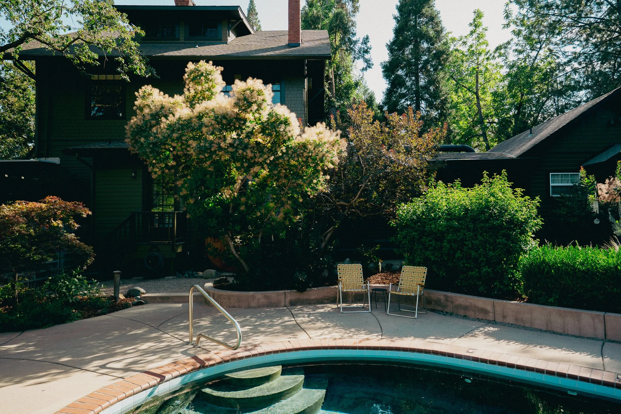 A backyard with a swimming pool in the foreground and a house with a steep roof in the background, surrounded by tall trees and lush greenery.