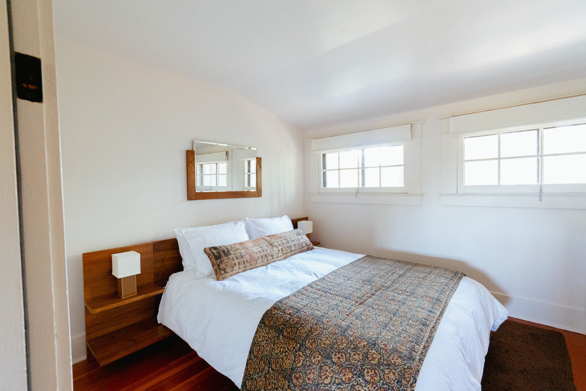 A cozy bedroom with a wooden bed frame, white bedding, decorative pillows, a patterned bed runner, large windows with white blinds, and a mirror on the wall.