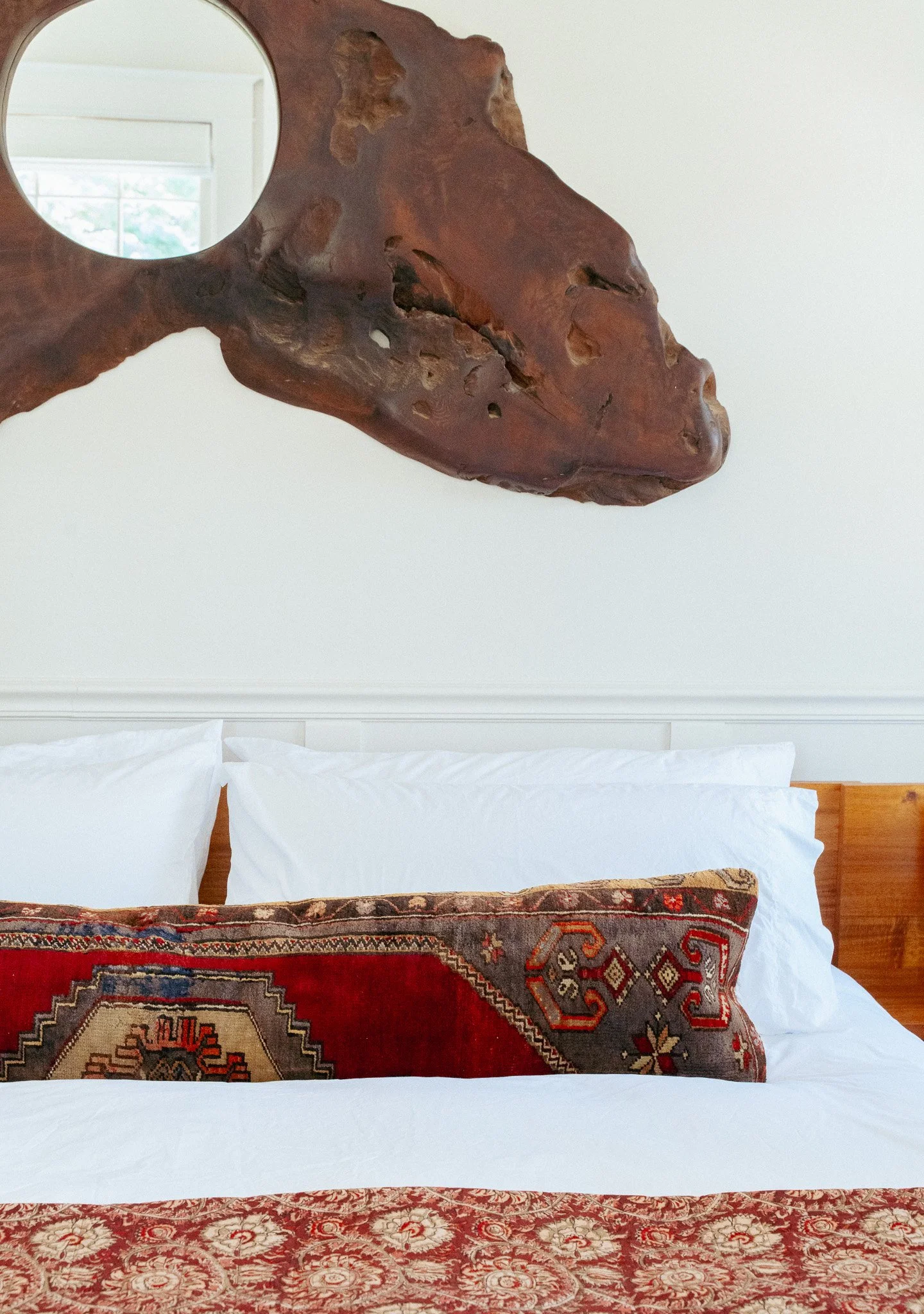 A close-up of a bed with white bedsheets, a decorative pillow with a colorful pattern, a red and gold patterned runner, and a wooden headboard, with a large wooden wall hanging resembling a dragon or animal head with a circular mirror as an eye.
