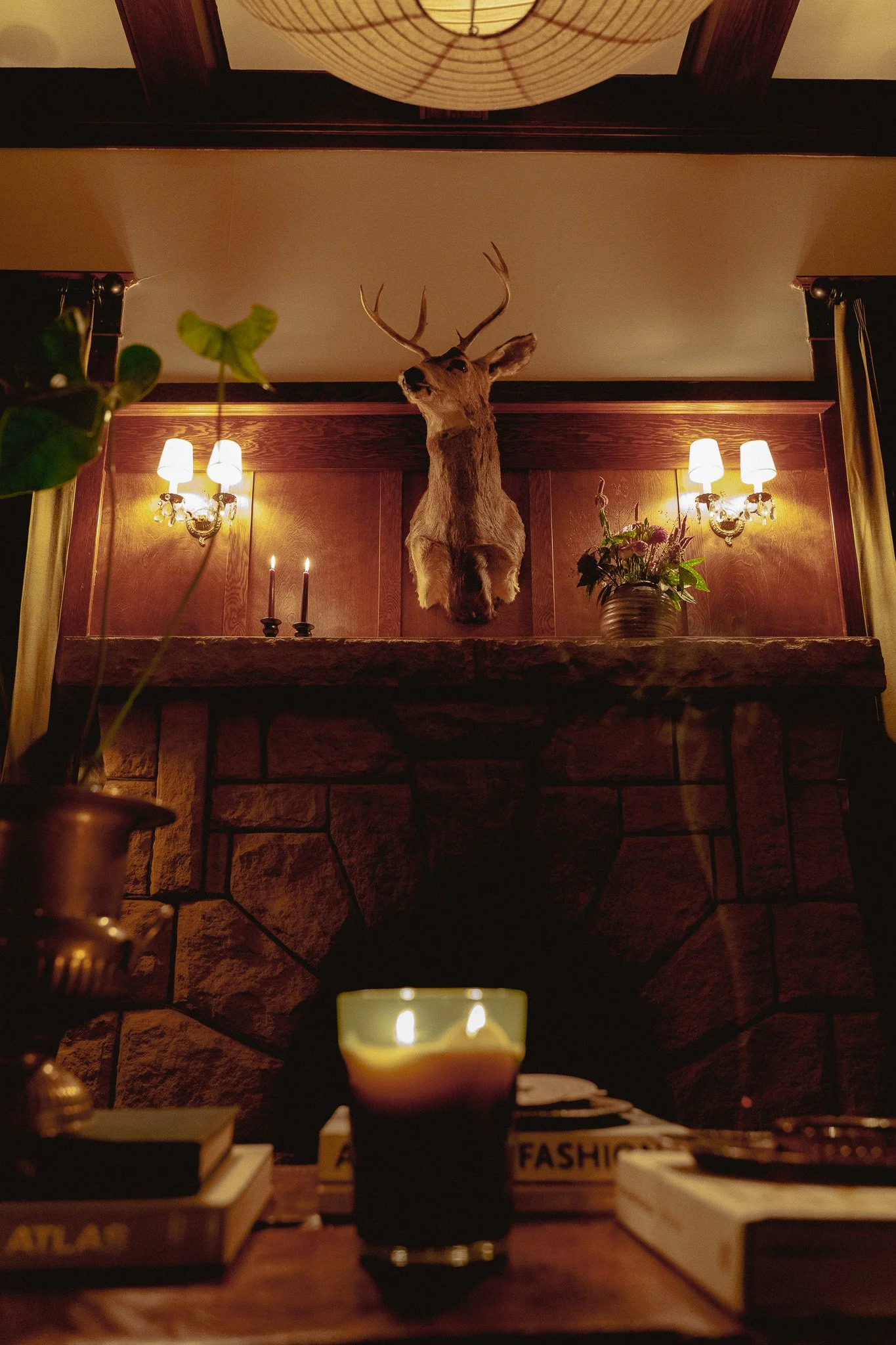 Wooden mantel with a mounted deer head with antlers, lit by two wall sconces, with candles and a flower vase on the shelf, and books and a candle in the foreground.