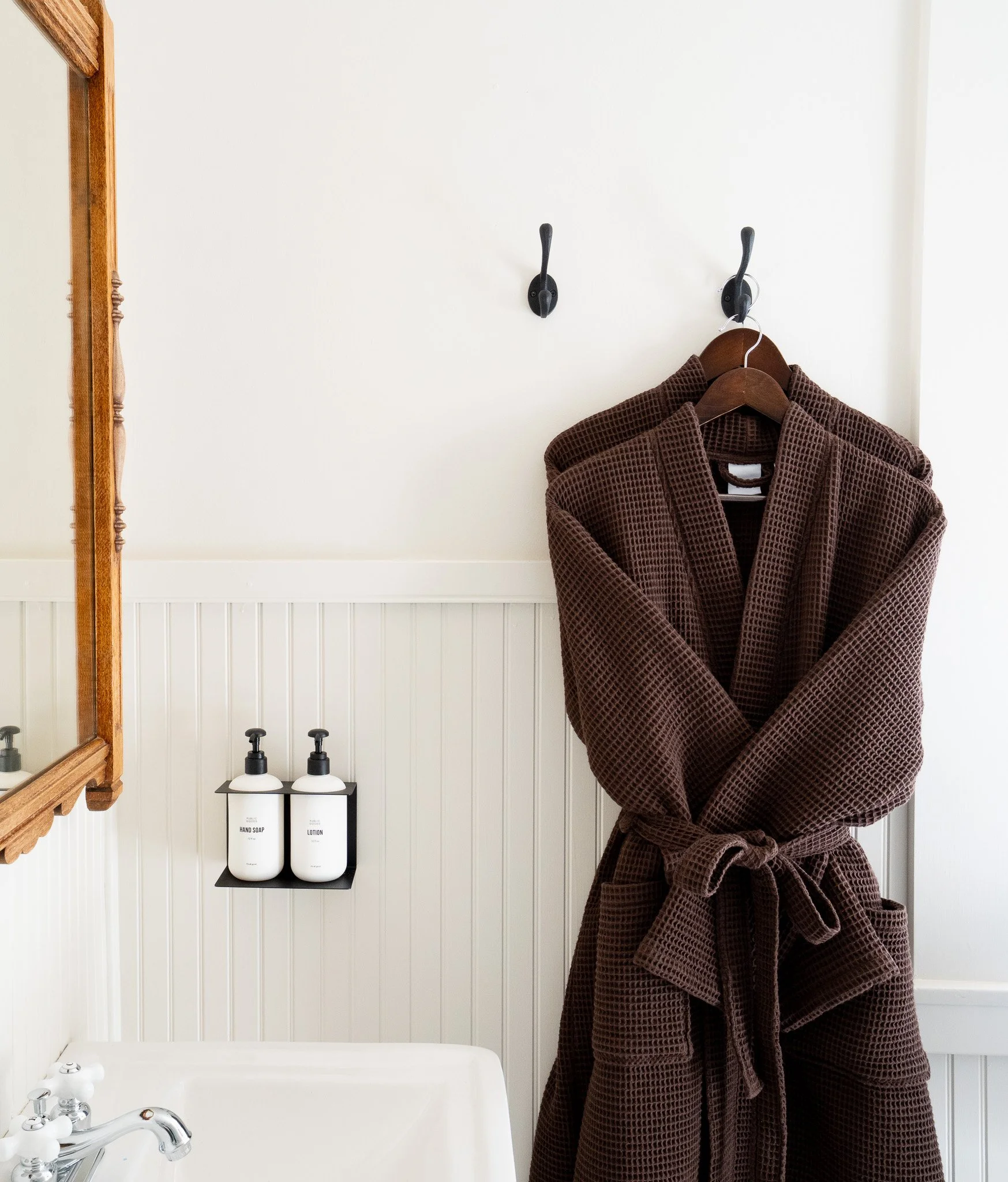 Bathroom corner with a brown bathrobe hanging on a black hook on a white wall, a wooden framed mirror on the left, two white bottles labeled 'Hand Soap' and 'Lotion' on a small black shelf, and a white bathtub with a silver faucet at the bottom.
