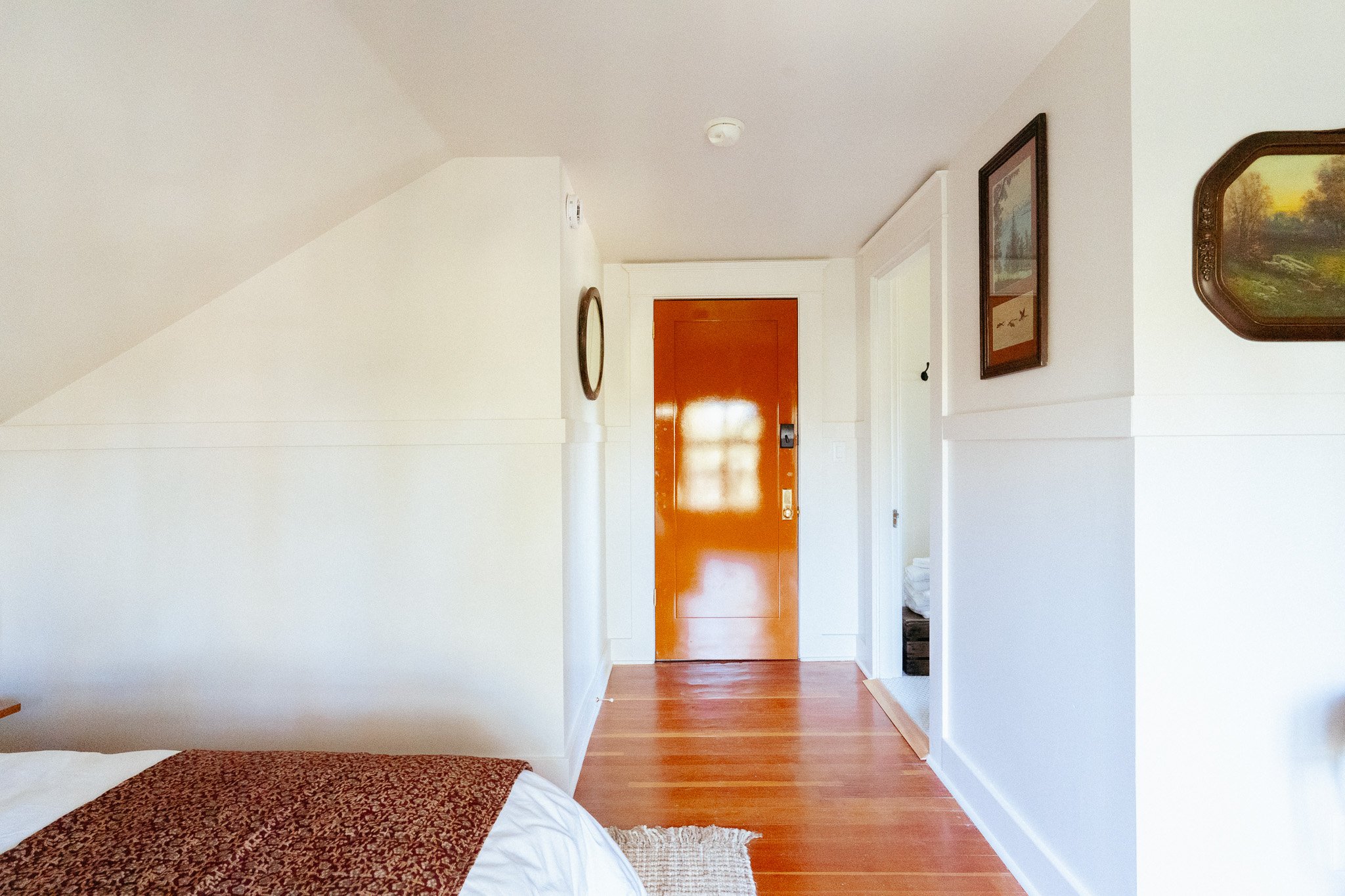 Interior of a room showing part of a bed with a brown patterned blanket, hardwood floors, white walls, a closed wooden door, framed artwork on the wall, and a small section of a staircase.