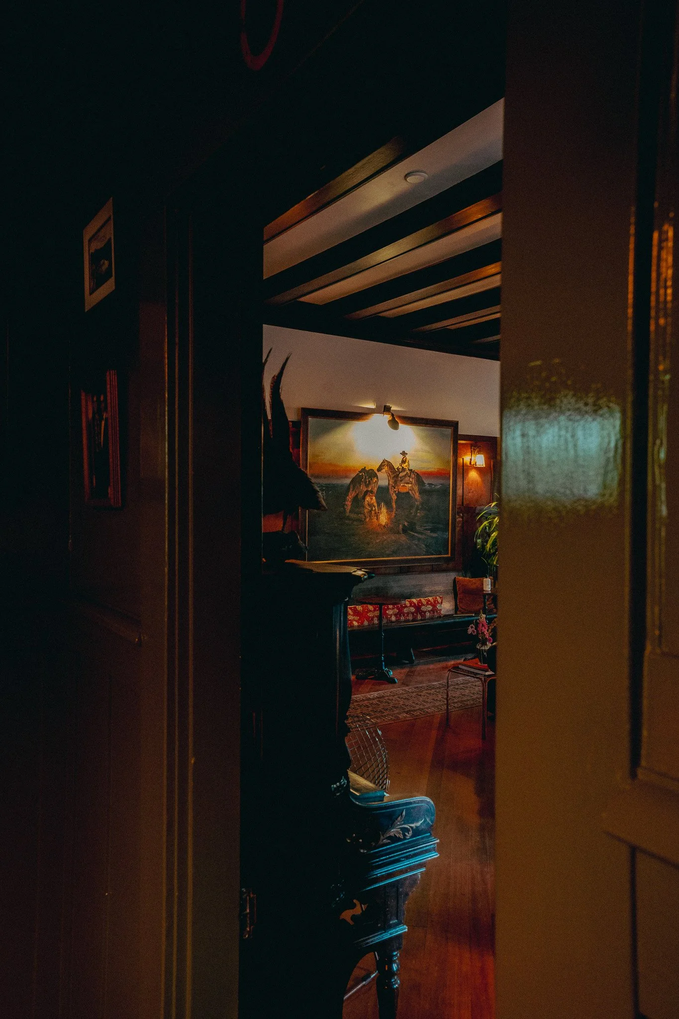 View of a cozy, dimly lit room with a large framed painting of horses on a beach, a wooden ceiling, and furniture including a chair and a table, seen through a doorway.