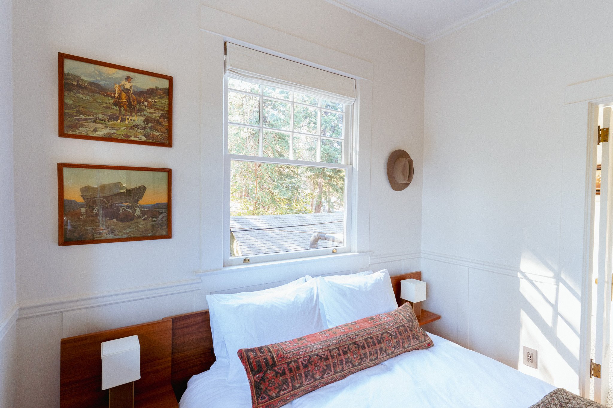 A bedroom with white walls, a wooden bed frame, white bedding, and decorative pillows. Two framed landscape paintings hang on the wall. A large window with a white shade lets in natural light, and a hat hangs on the wall near the window.