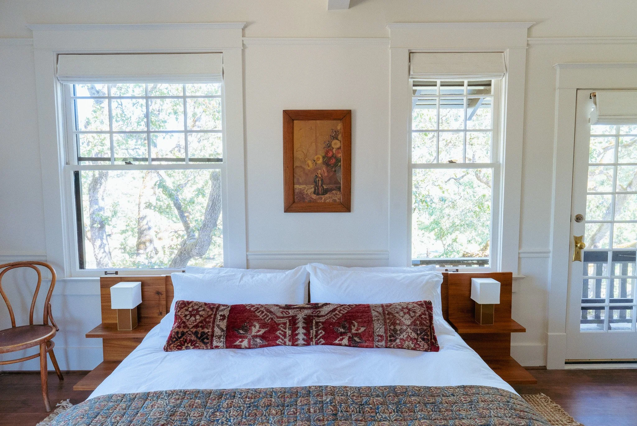 A bedroom with two large windows behind a bed. The bed has white linens and a decorative pillow. There is a framed painting of flowers on the wall between the windows. To the right, there is a door with glass panes and a brass handle. A wooden chair is partially visible on the left side of the image.
