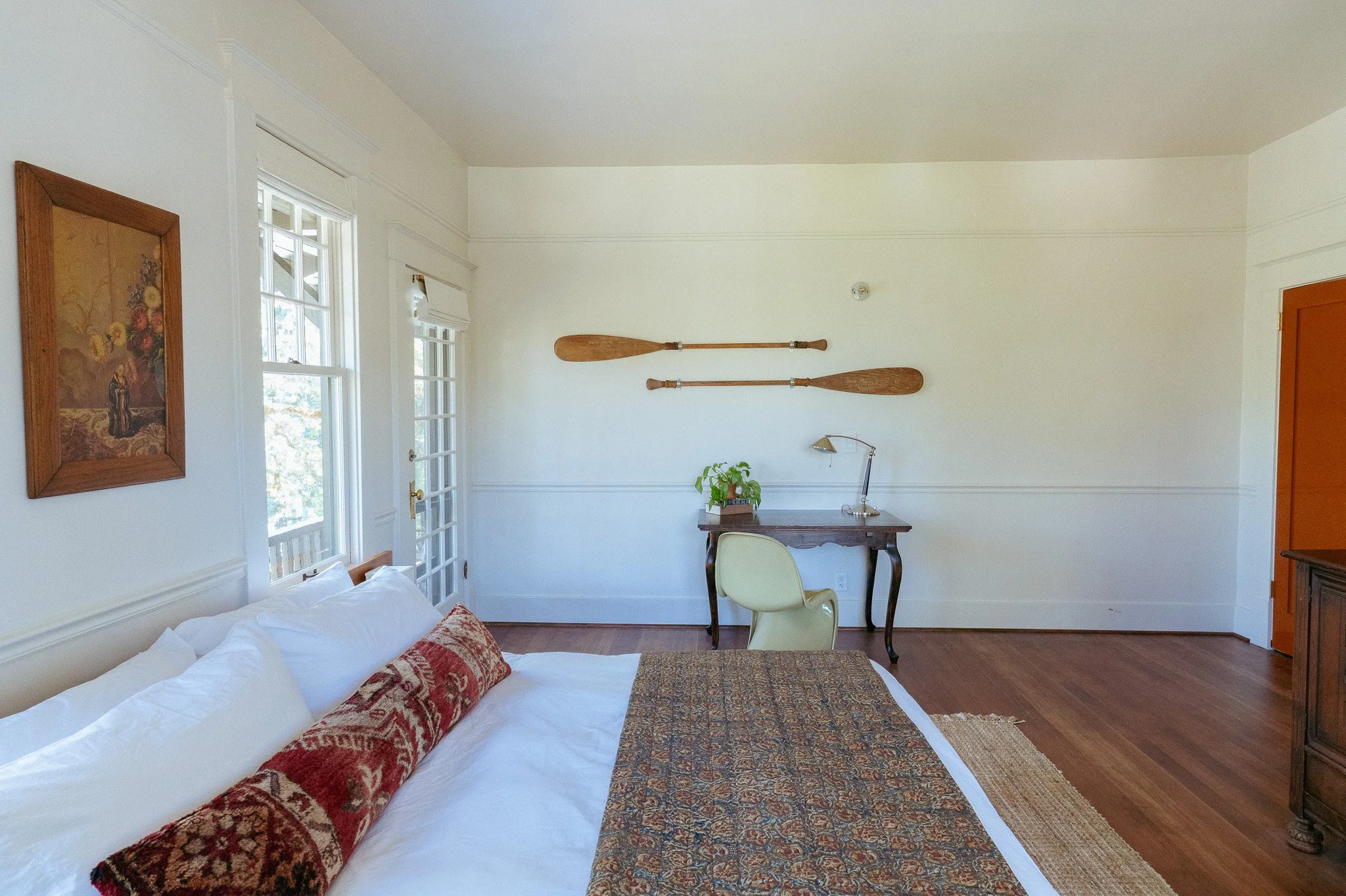 A corner of a bedroom with white walls, a bed with white bedding and decorative pillows, a wooden desk with a chair, potted plant, and desk lamp, two long wooden paddles mounted on the wall, a framed painting, and windows with white trim.