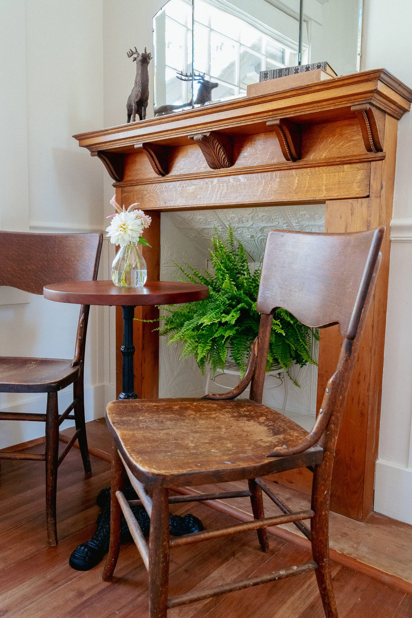 Wooden chairs around a small round table with a glass vase containing white and pink flowers, a green fern plant underneath, and a wooden mantle shelf with decorative objects including a reindeer figurine and a mirror reflecting a window.
