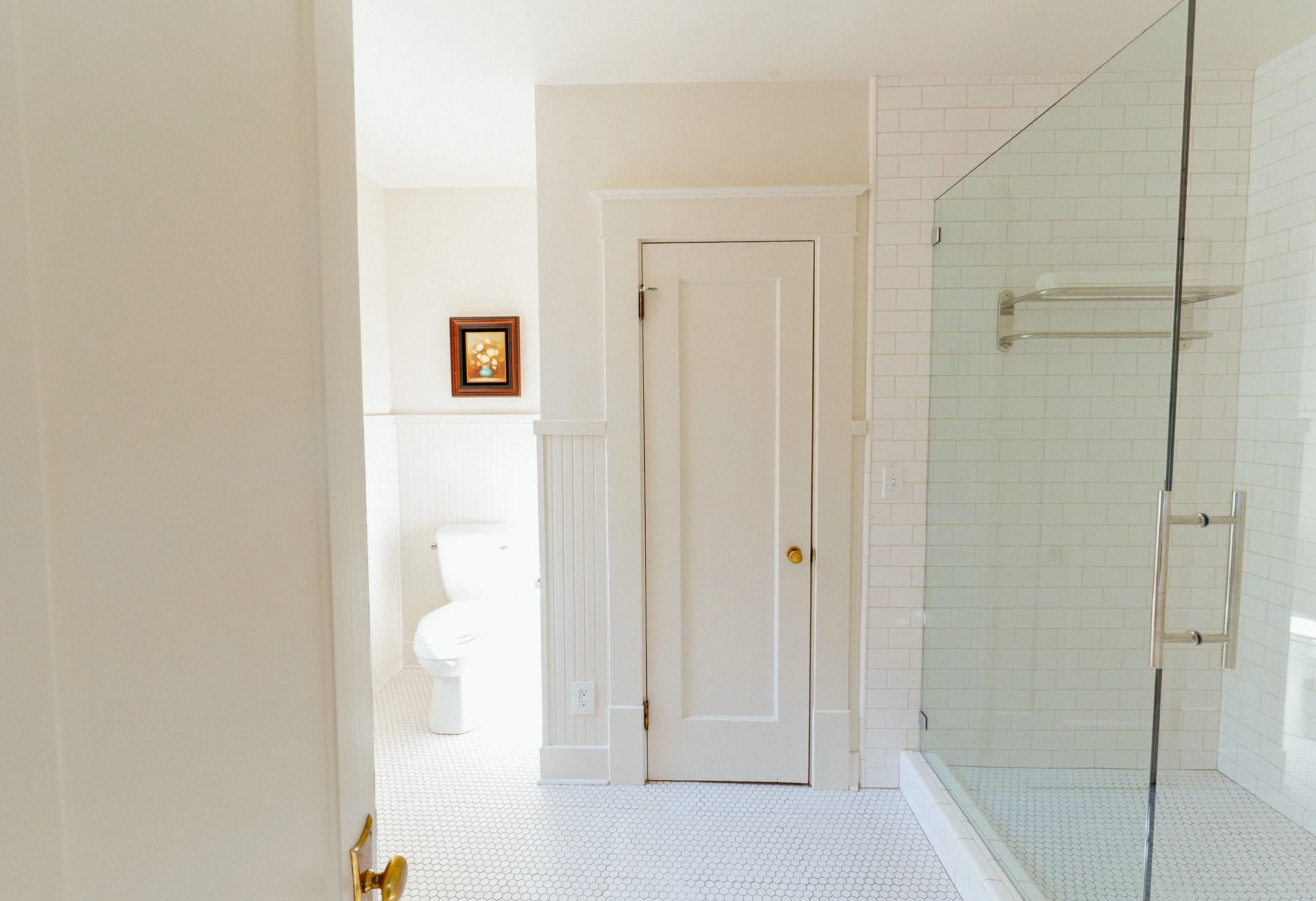 Interior view of a bathroom with a glass shower enclosure, white tiled walls, a toilet, a door, and wall art.