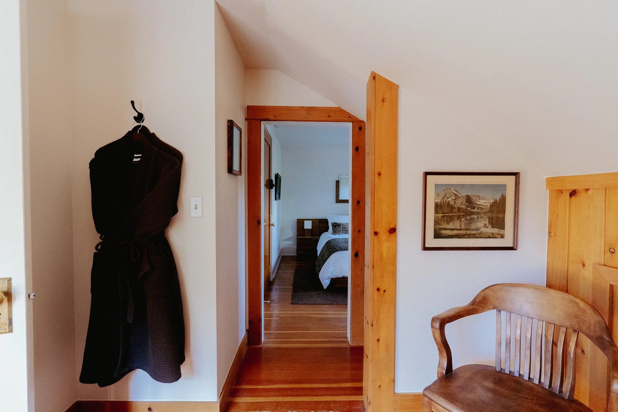 View of a bedroom through an open doorway, with a bed, framed artwork, wooden trim, and a chair visible, with a coat hanging on a hook on the wall.