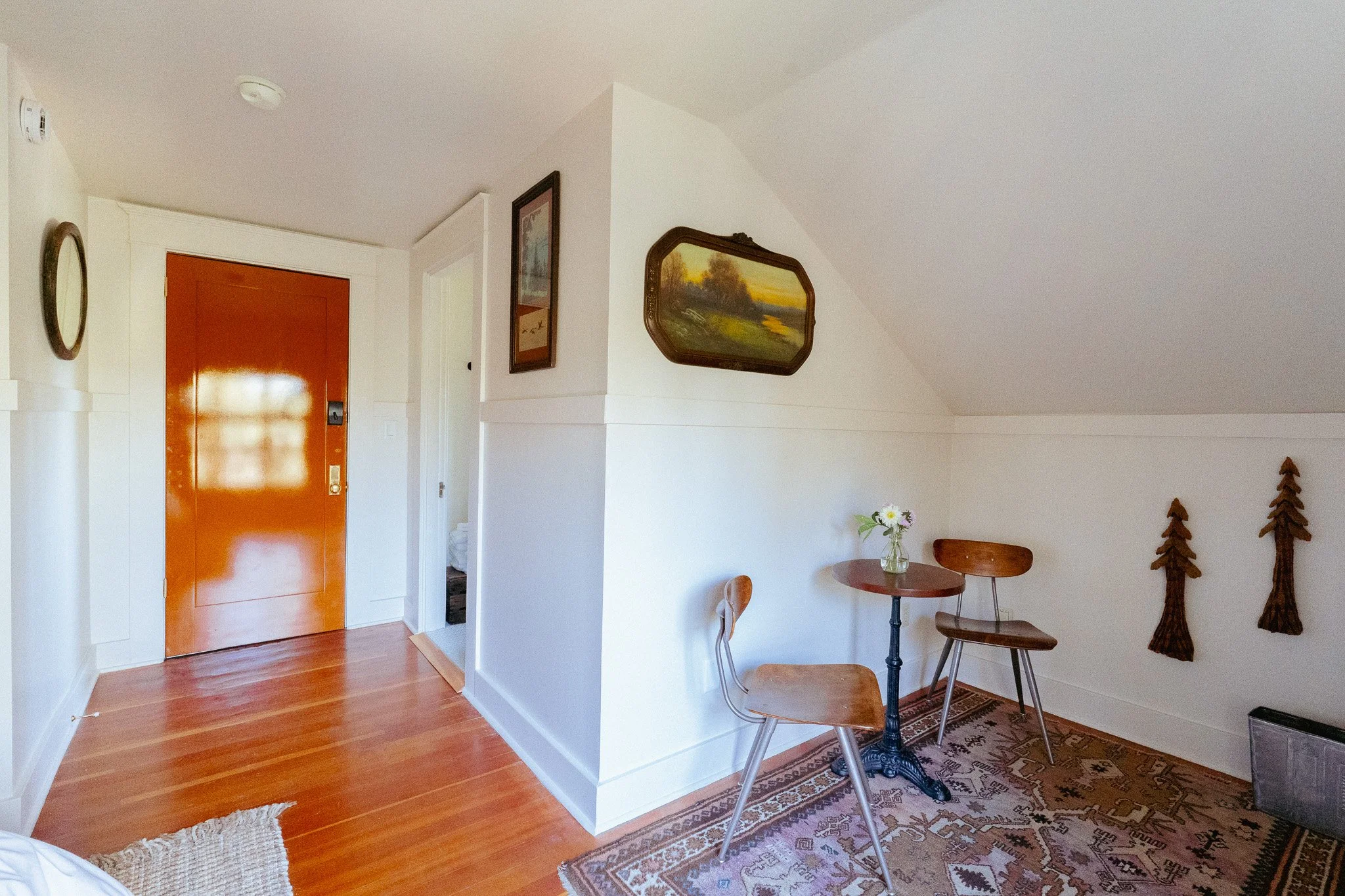 Interior view of a room with a wooden floor, white walls, and decorated with framed landscape paintings. Visible furniture includes a small round table with two wooden chairs, a decorative area rug, and a vase with flowers. There are also wooden ornaments on the right side and a partially open doorway with a bright orange door.