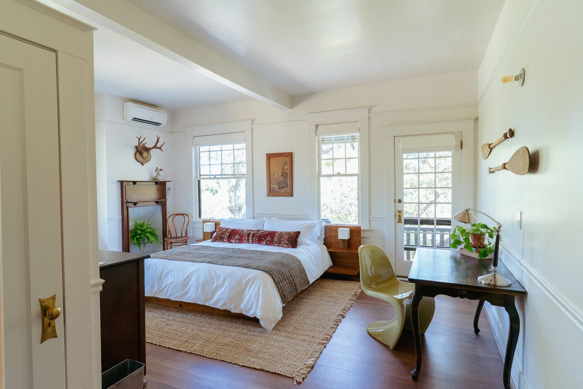 Bright bedroom with white walls, large windows, a bed with white and patterned bedding, a small wooden desk with a vintage lamp, a yellow chair, and plants. Decor includes a deer head mount and framed art.