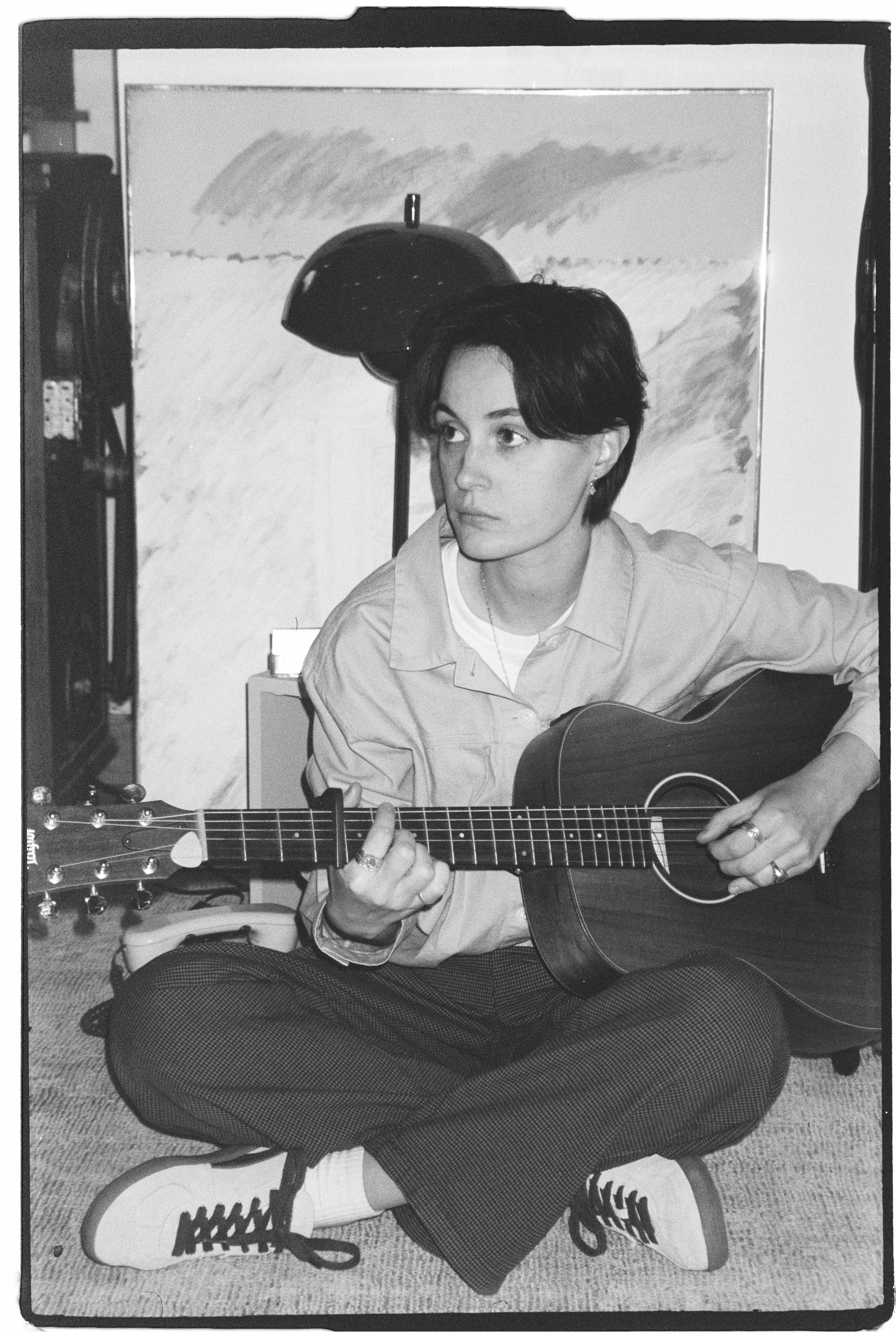 Photo de Rosemarie Gauthier. Une femme assise par terre, jouant de la guitare acoustique, portant des vêtements décontractés, des baskets Nike blanches, et écoute de la musique dans une pièce avec un tableau en arrière-plan.