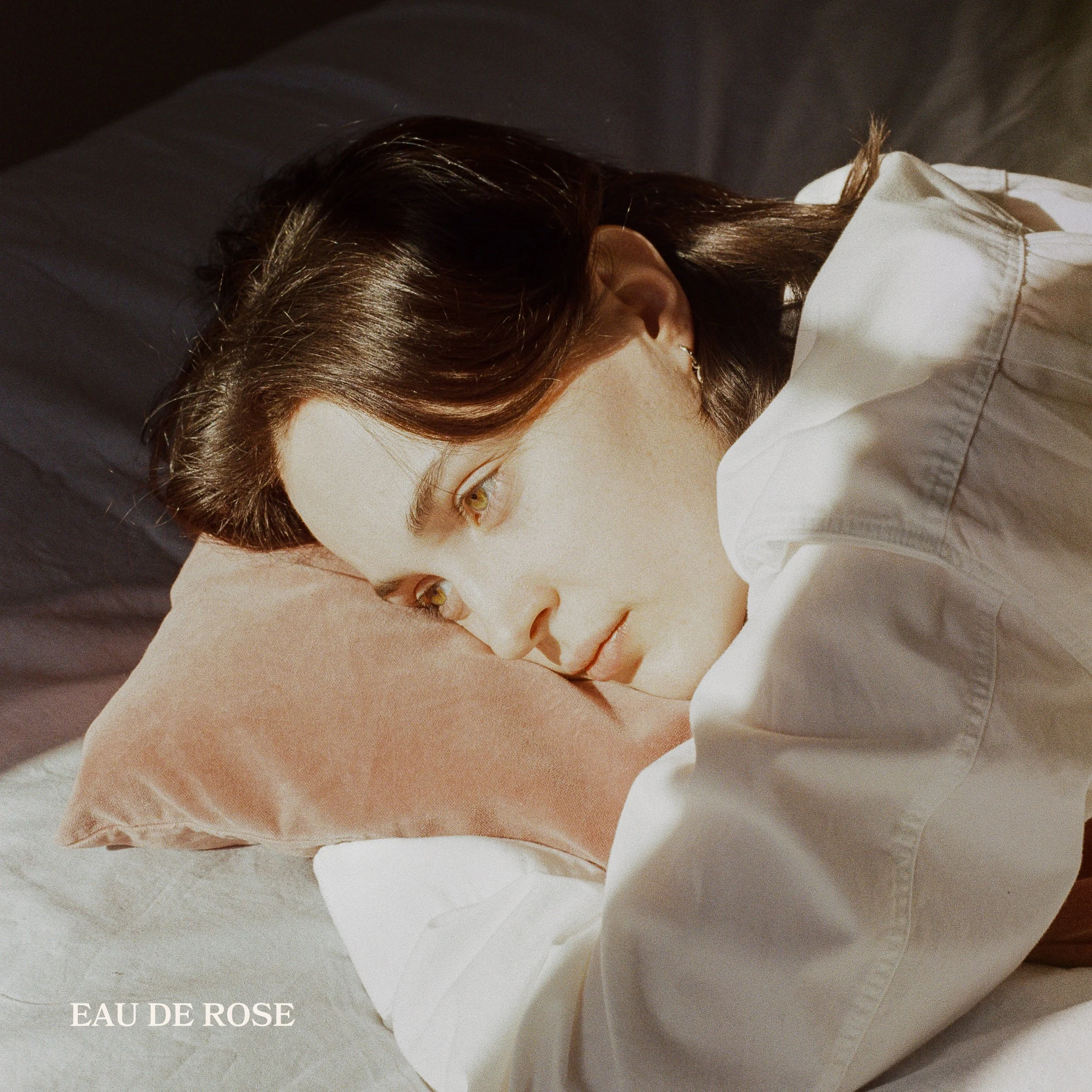 Pochette du EP Eau de rose de Rosemarie Gauthier. Femme avec des cheveux bruns longs portant un chemisier blanc, vue de côté, exposant partiellement son visage et son oreille, avec le texte 'EAU DE ROSE' en bas à gauche.