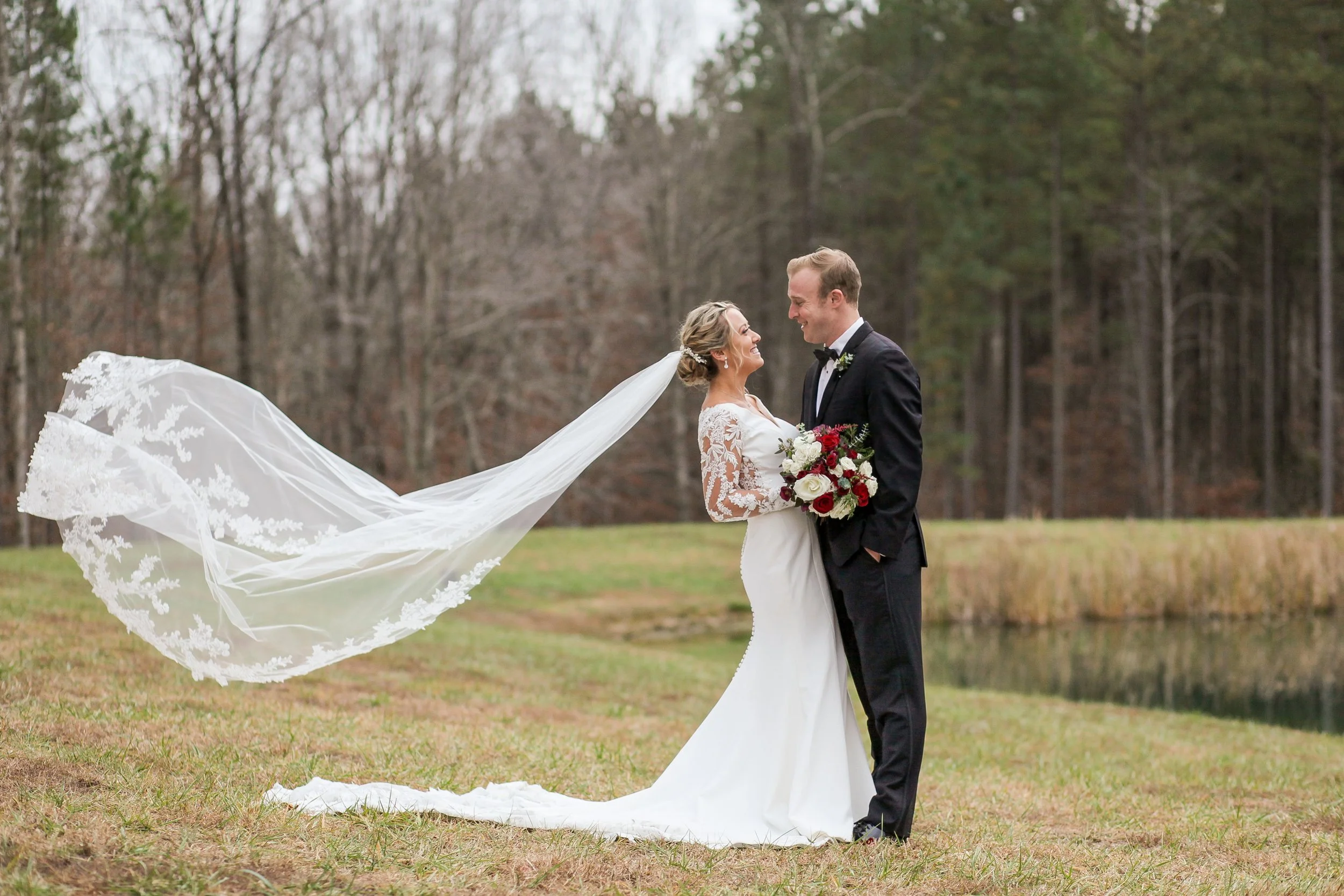 A bride and groom standing outdoors in a grassy area with trees in the background, smiling and looking at each other, the bride holding a bouquet of white and red flowers, and her long veil flowing in the wind.