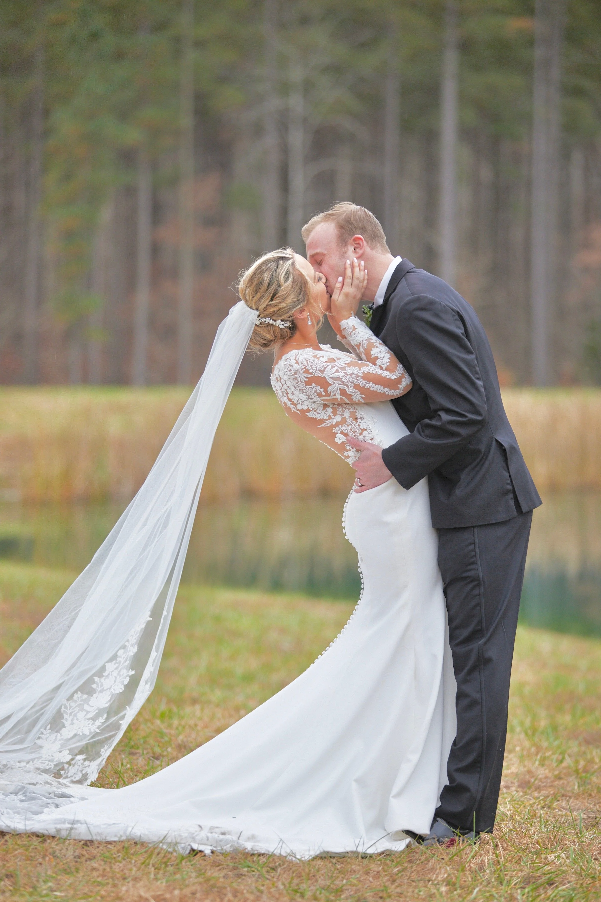A bride and groom share a kiss outdoors during their wedding, with the bride's veil flowing on the ground and the groom wearing a black suit on a grassy area with trees in the background.