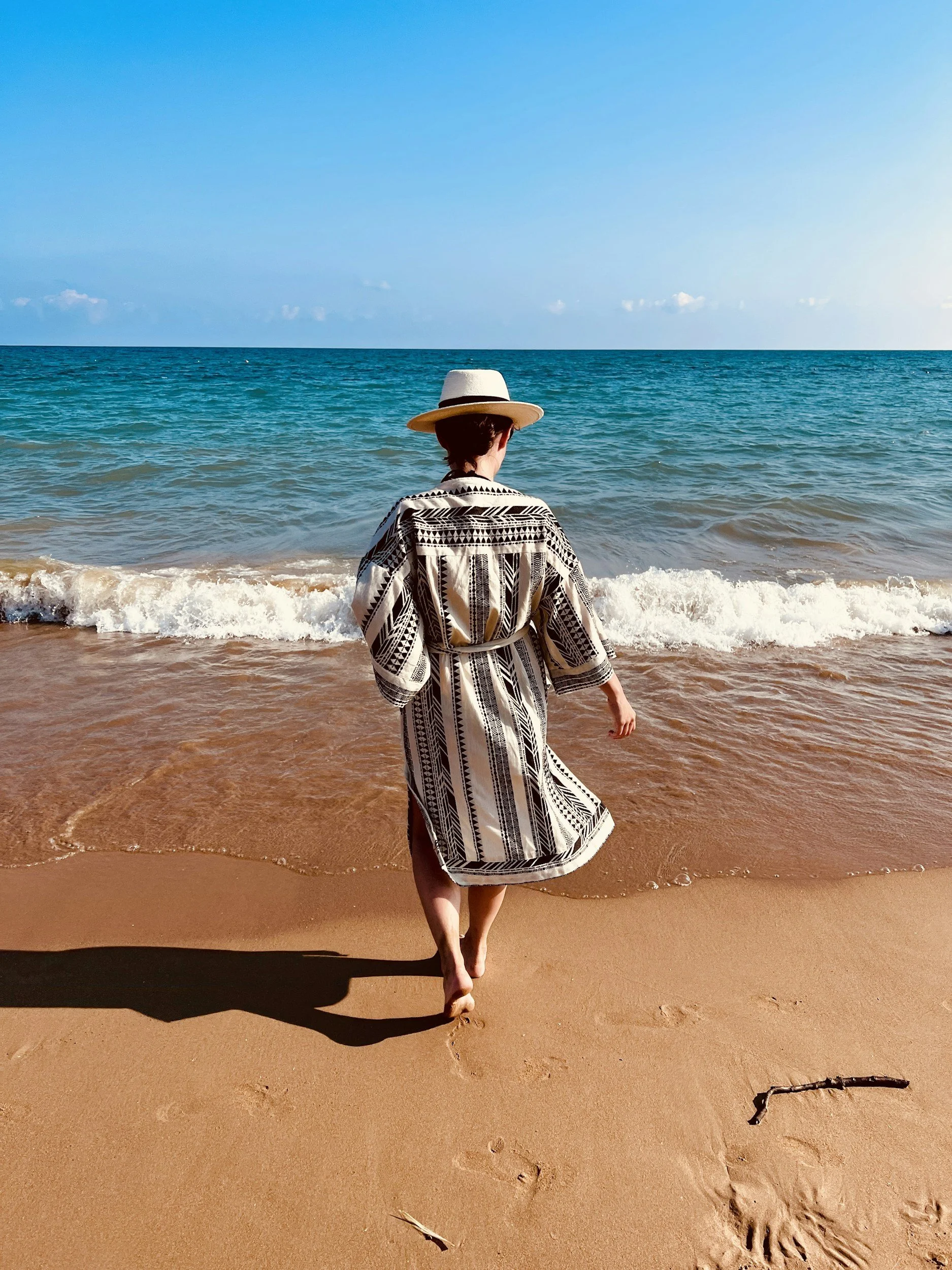 A person wearing a black and white patterned robe and a white hat walking along the sandy beach toward the ocean, with waves approaching the shore and footprints in the sand.