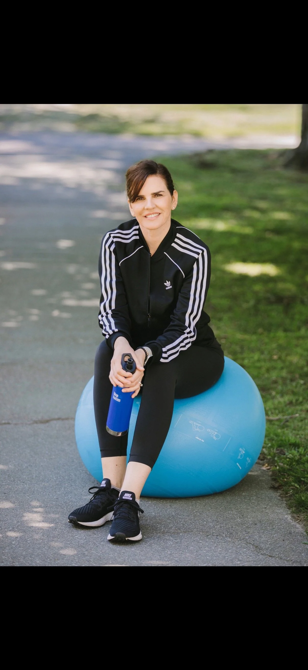 Woman in black athletic jacket with white stripes sitting on a blue exercise ball, holding a water bottle, smiling outdoors on a paved path with green grass and trees in the background.