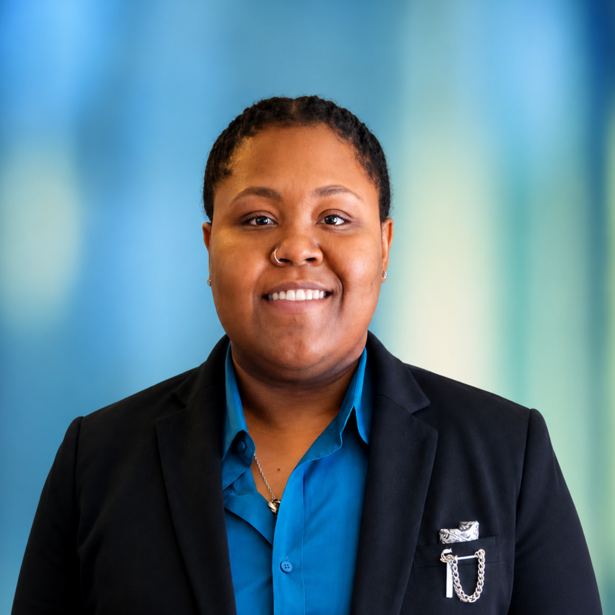 A young person with short curly hair and a nose ring, wearing a black blazer over a blue shirt, smiling in front of a colorful blurred background.
