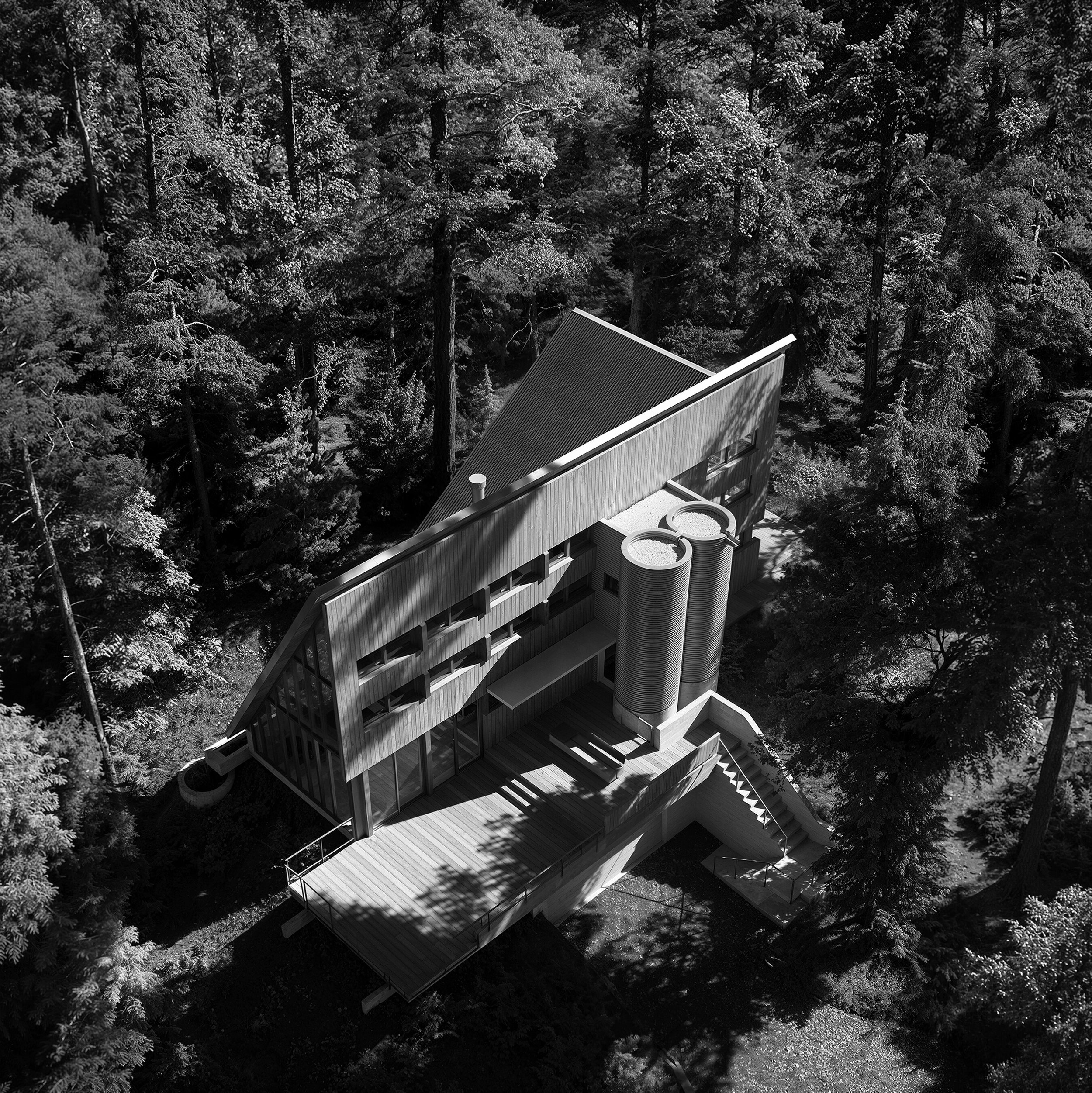 Aerial view showing the Italic A house, roof geometry, deck, and cylindrical towers within the forest