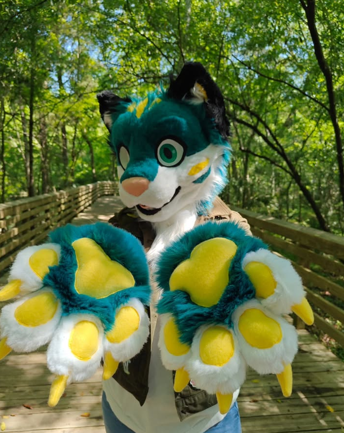 Person in a colorful fursuit with teal, white, black, and yellow features, wearing large paw gloves with yellow pads, standing on a wooden bridge in a green forest.