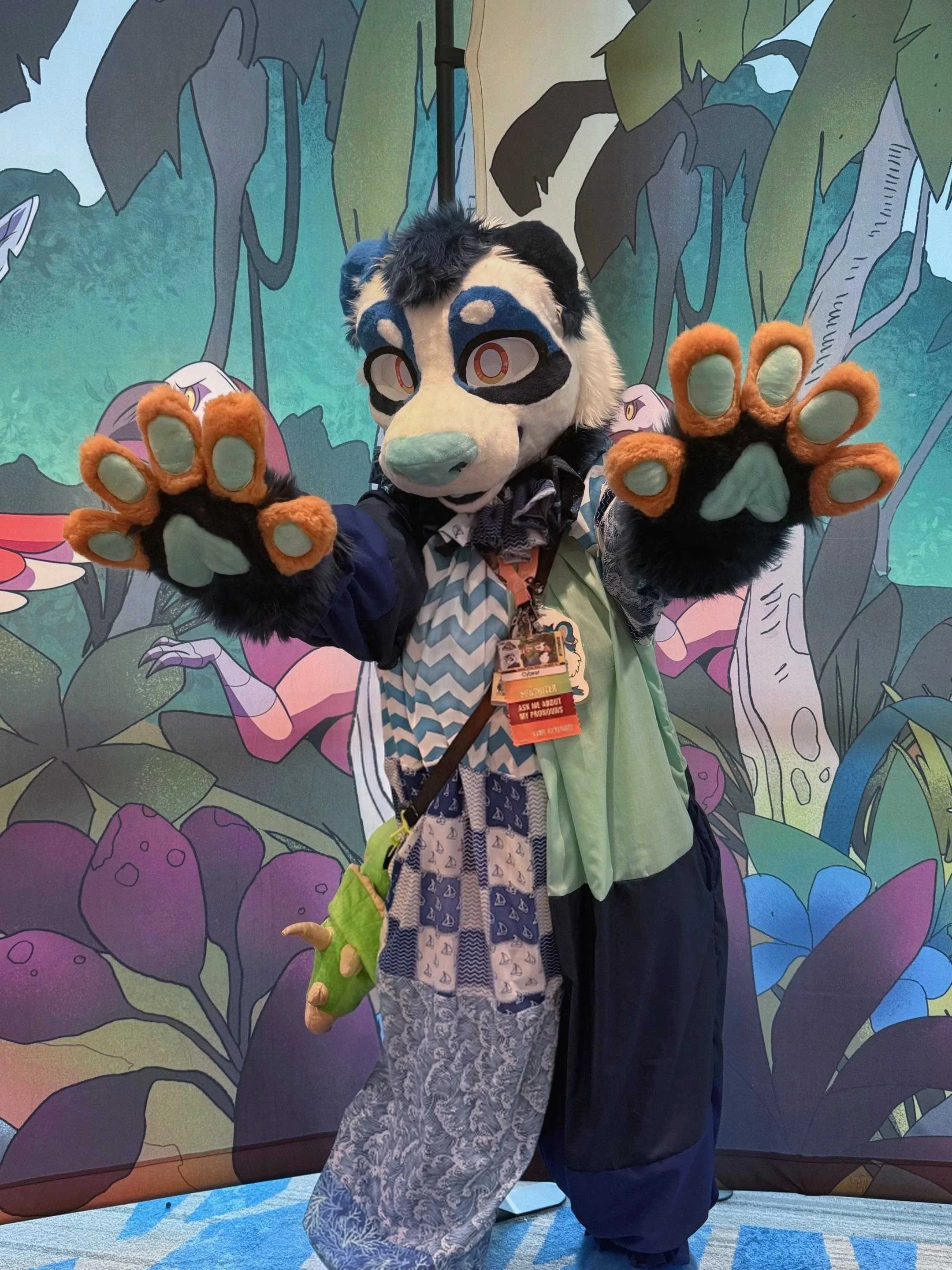 Person in a furry animal costume, resembling a panda or raccoon, wearing a black, white, and blue outfit with a floral pattern, holding up their paws with orange and mint green gloves, standing in front of a colorful illustrated jungle background.