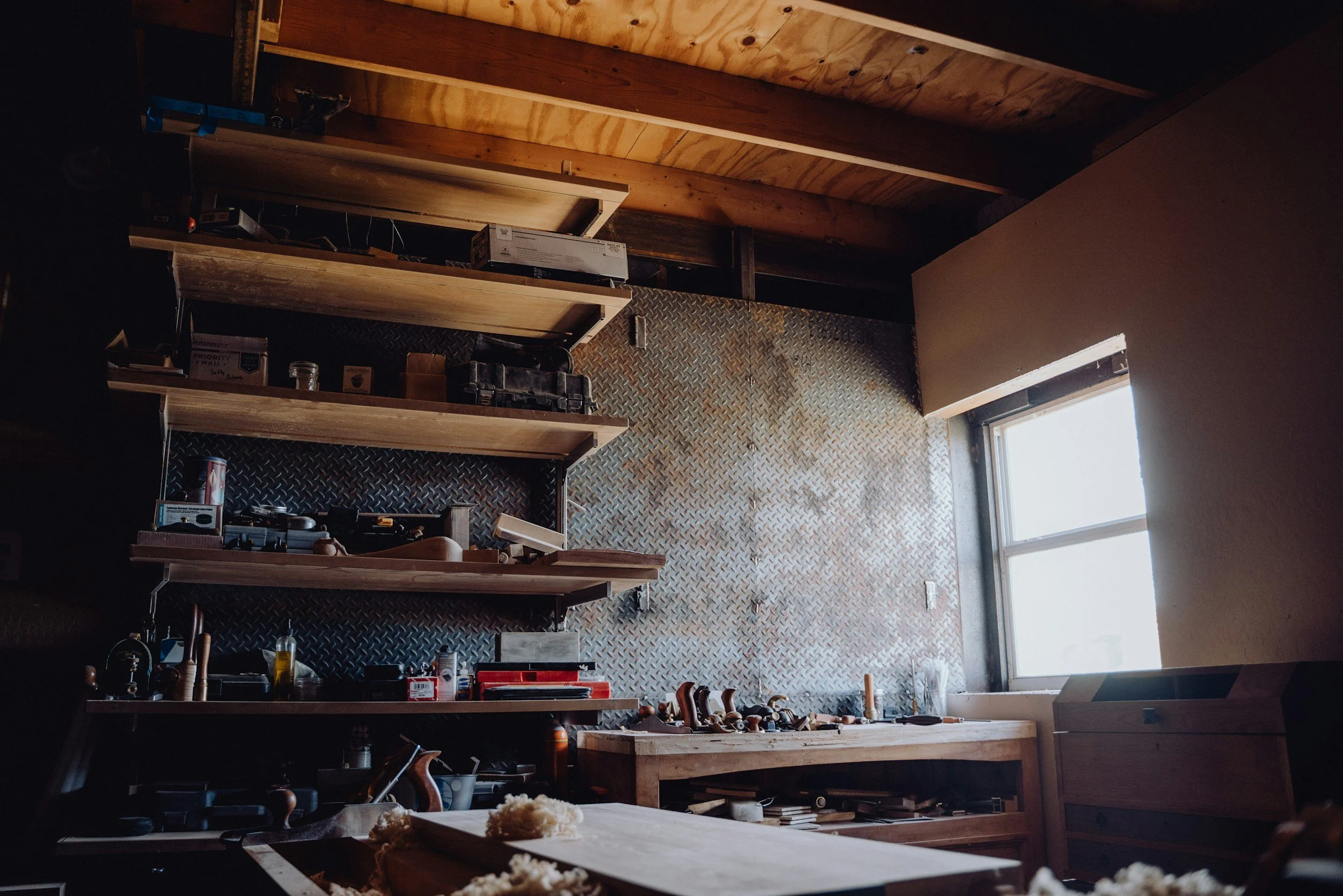 A woodworking workshop with wooden shelves and a workbench near a window, cluttered with tools and supplies.