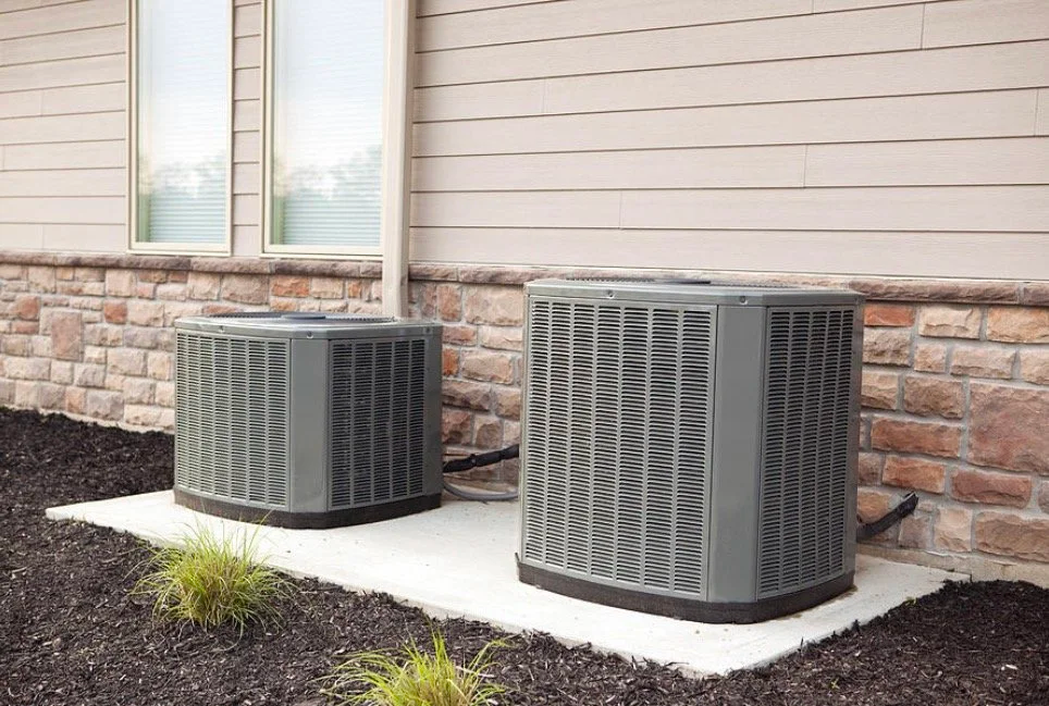 Two outdoor air conditioning units installed on a concrete slab next to a house with a brick and siding exterior.
