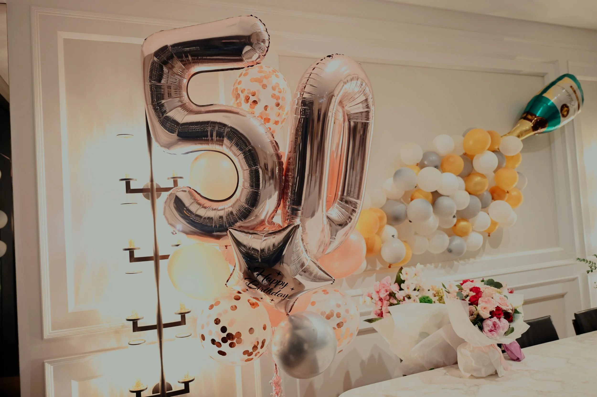 Birthday celebration decorations with large silver balloons spelling out the number 30, a cluster of balloons in white, silver, and gold, and a bouquet of pink and red flowers on a decorated table in a room with beige walls and wall sconces.