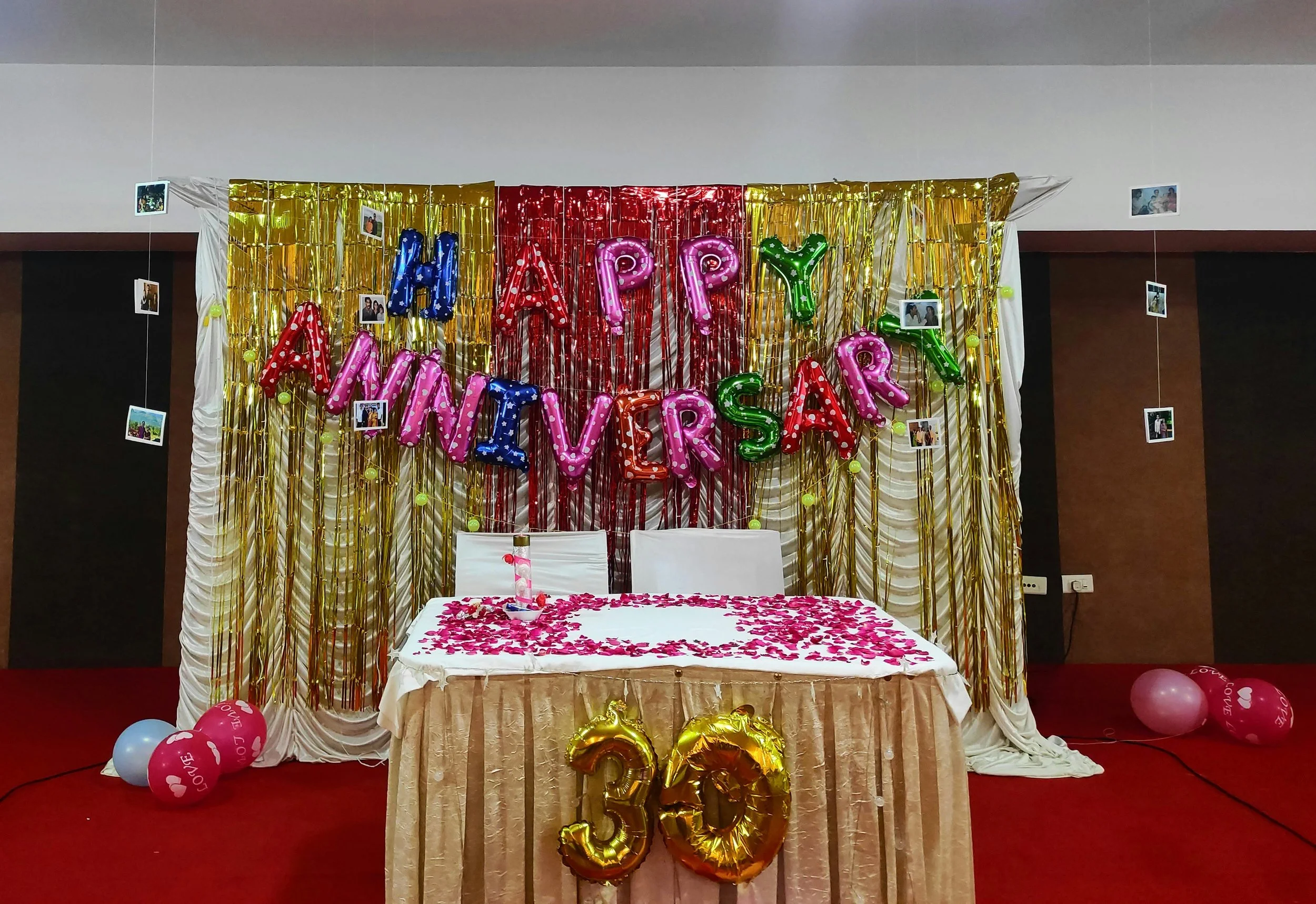Decorated party setup with a backdrop that says 'Happy Anniversary Love' in colorful balloons, photos hanging on strings, a table with pink rose petals and balloons, and gold balloon numbers '30' at the front.