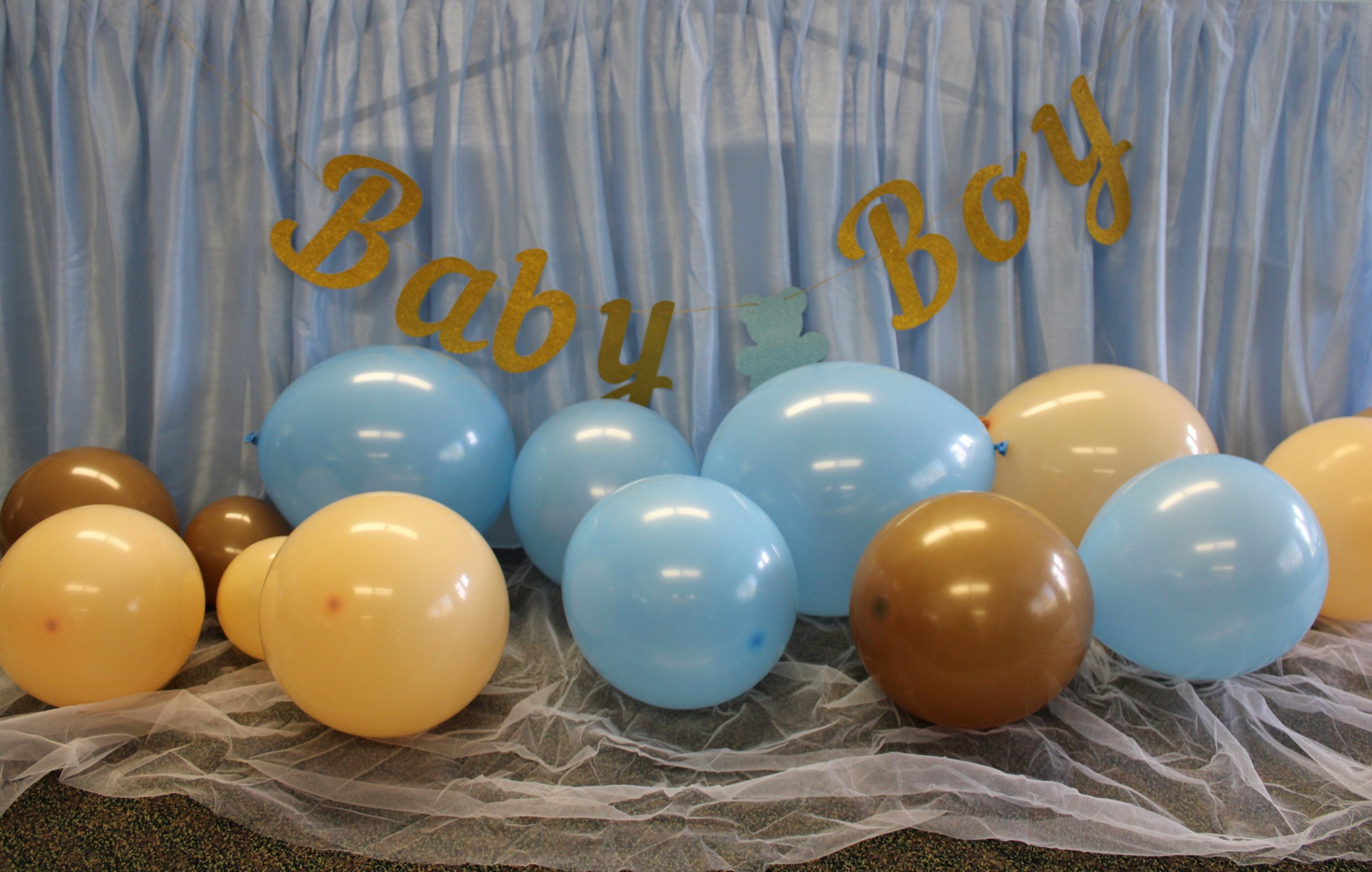 A colorful display with pastel blue and yellow balloons at the bottom, a blue curtain backdrop, and a gold glitter sign that reads "Baby Boy" with an angel wing decoration.