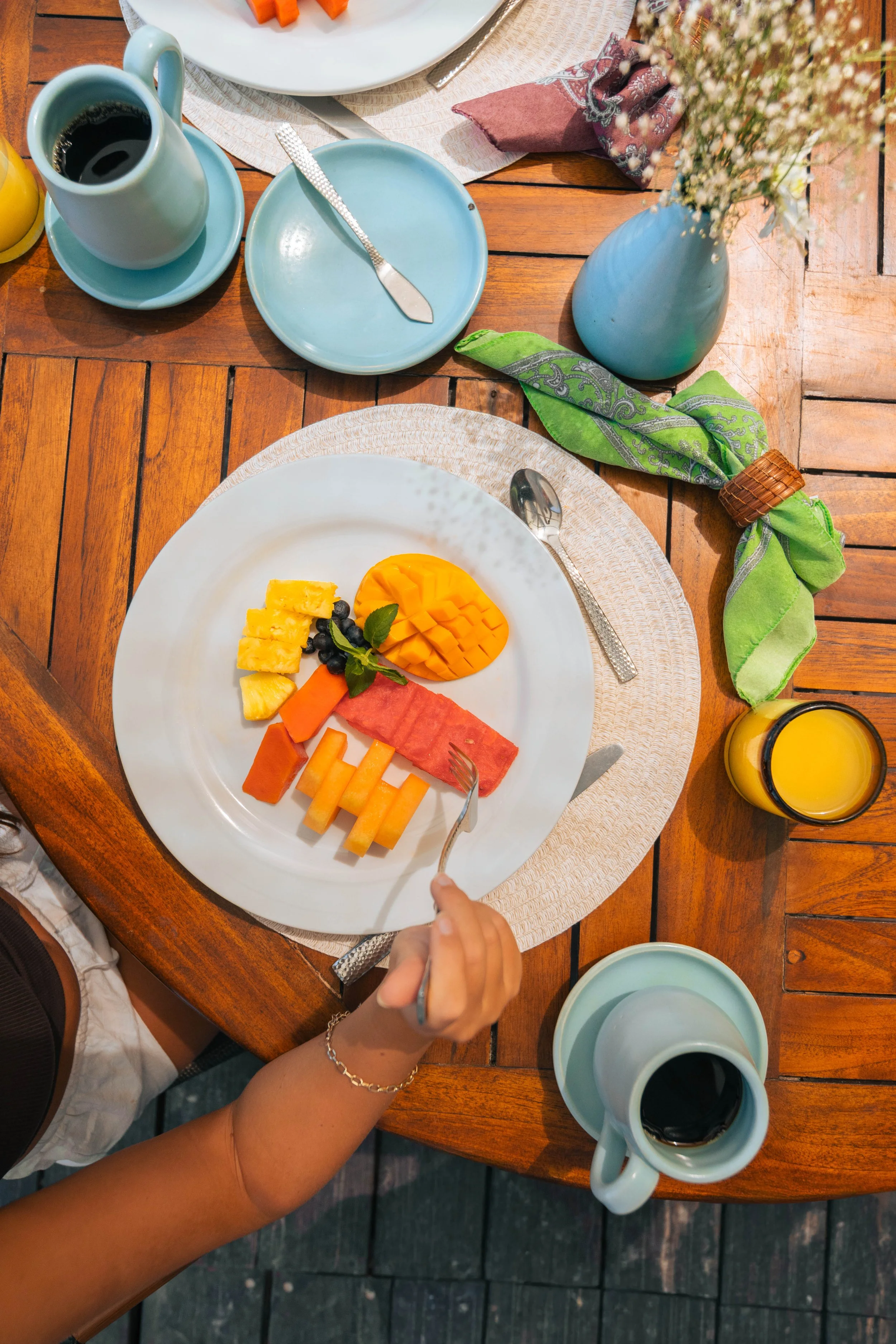 Plato con fruta como piña, mango, sandía, y arándanos, con una bebida y café en la mesa de madera.