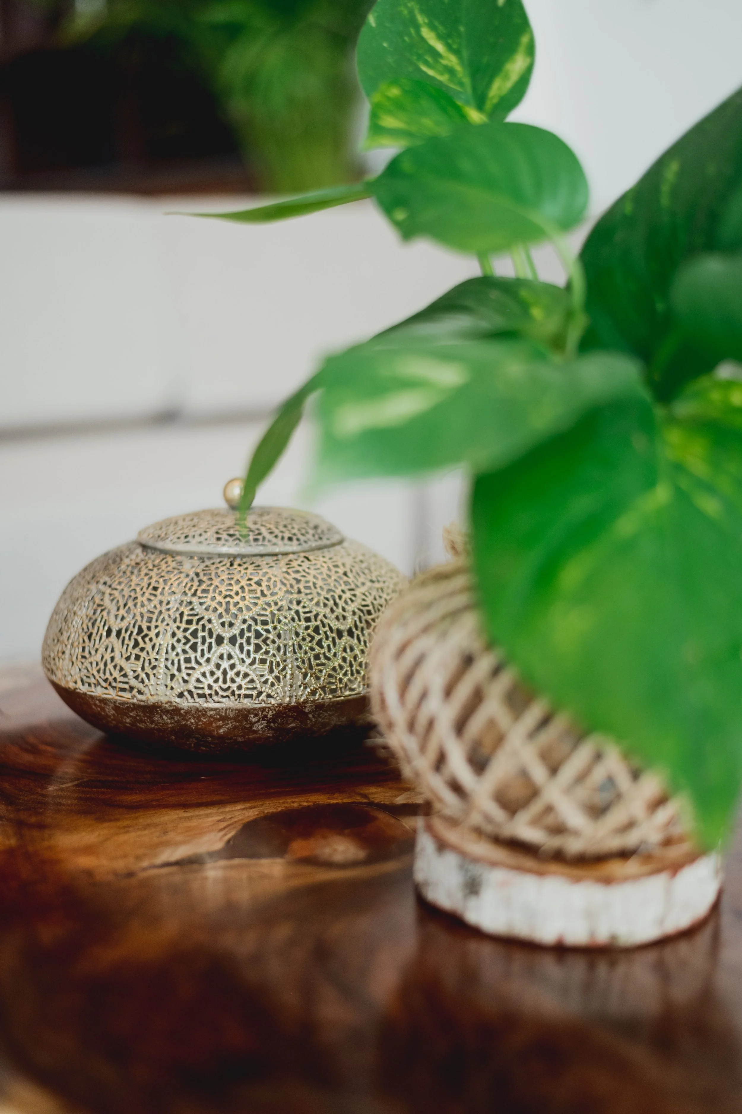 Decorativa lámpara de cerámica de color beige con patrones entrecruzados y base marrón, rodeada de plantas verdes en cestas de mimbre, sobre una mesa de madera.
