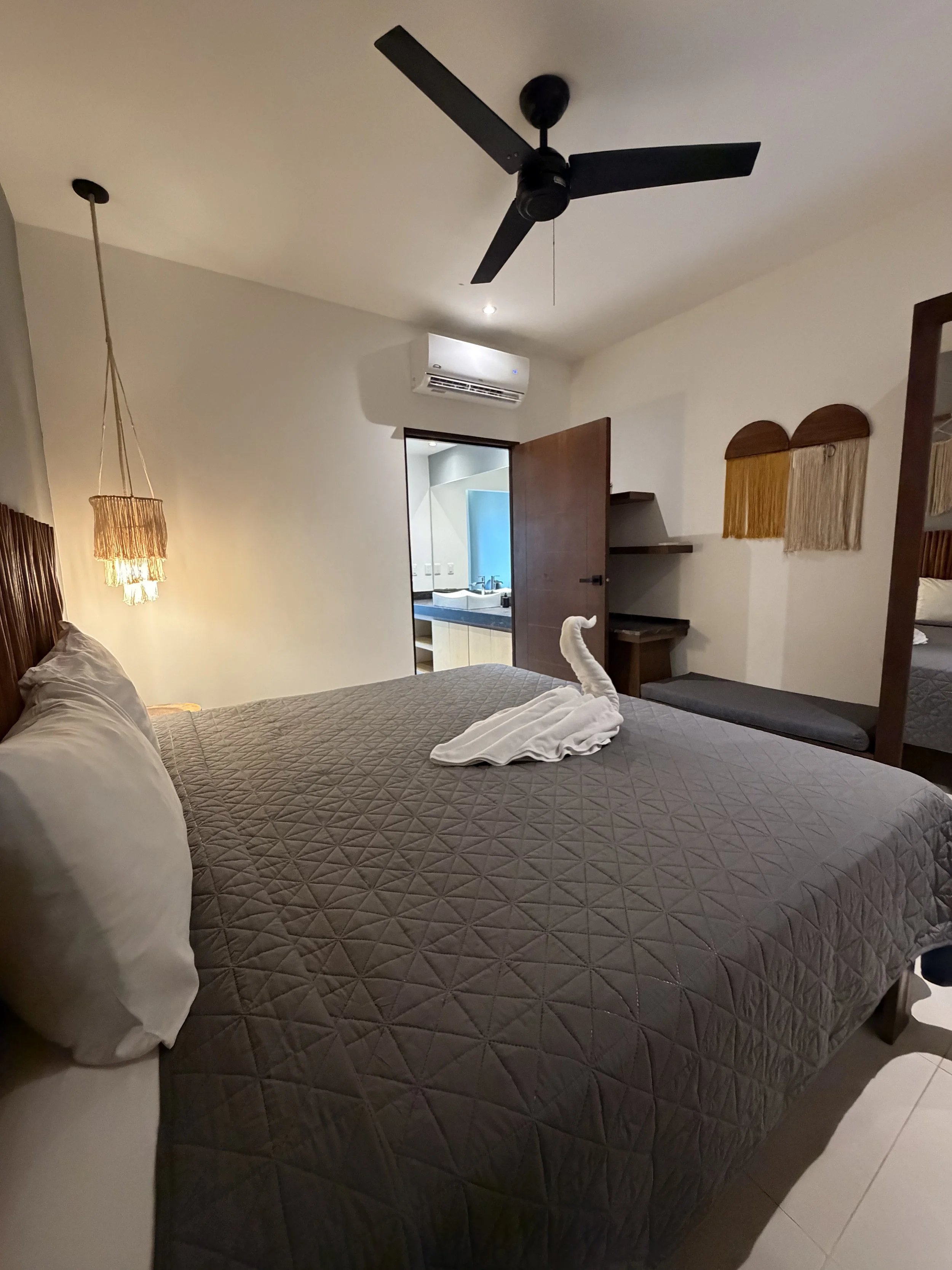 Habitación con cama de sábanas grises y almohadas blancas. Un toalla doblada en forma de cisne en la cama. Ventilador de techo negro. Lámpara de pared con pantalla de mimbre. Puerta abierta que lleva a un baño con lavabo y espejo.