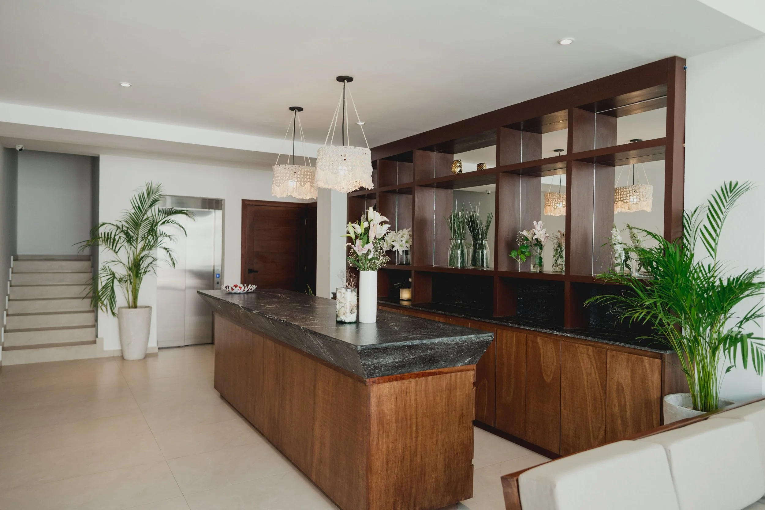 Interiores de una cocina moderna con mesón de granito oscuro, estanterías de madera, plantas decorativas, y lámparas colgantes con cortinas de ganchillo.
