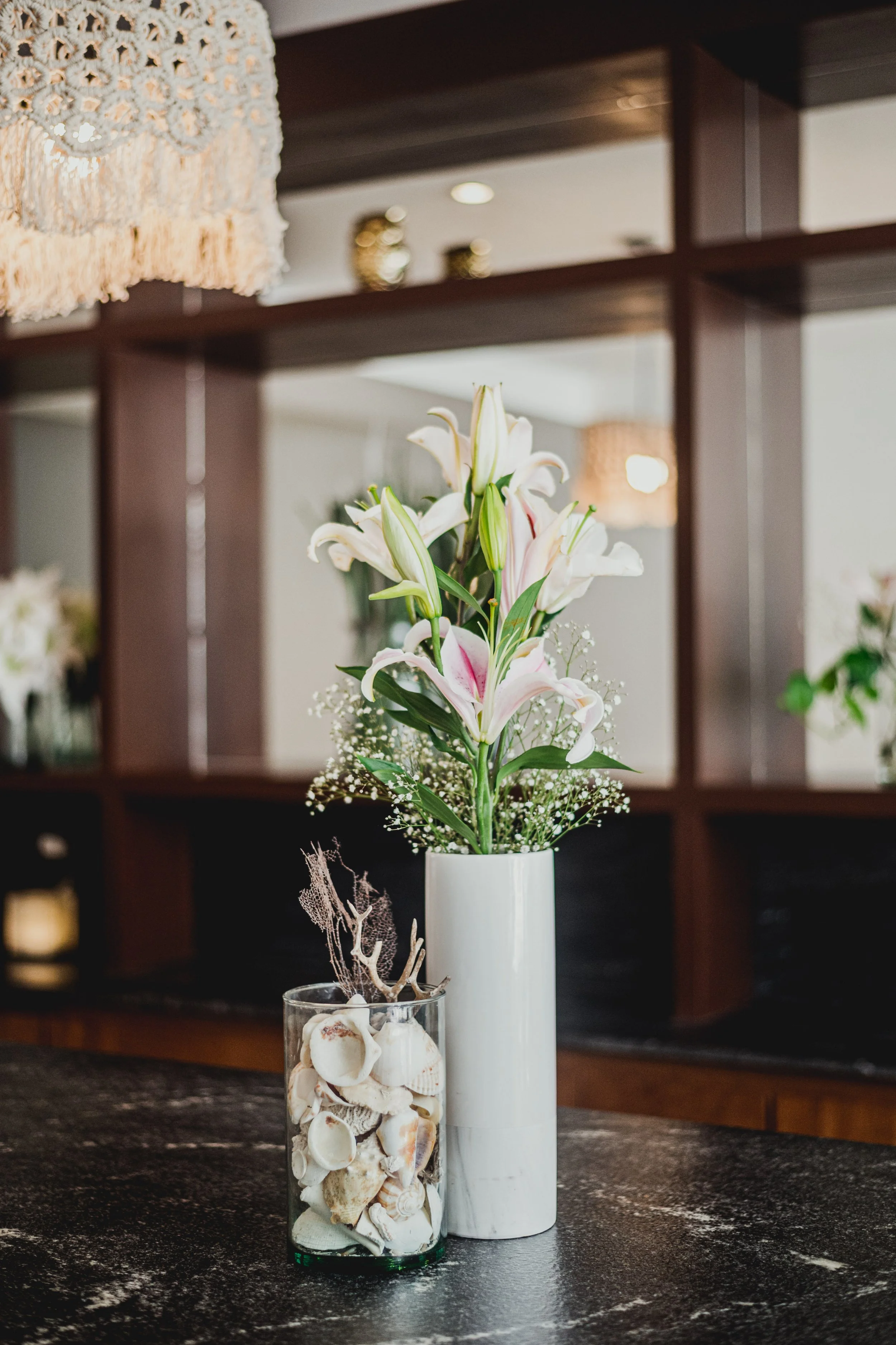 Un florero blanco con flores blancas y rosadas en una mesa de cocina, con una copa llena de conchitas y decoraciones de maderas, y en el fondo estanterías de madera en un ambiente rústico.