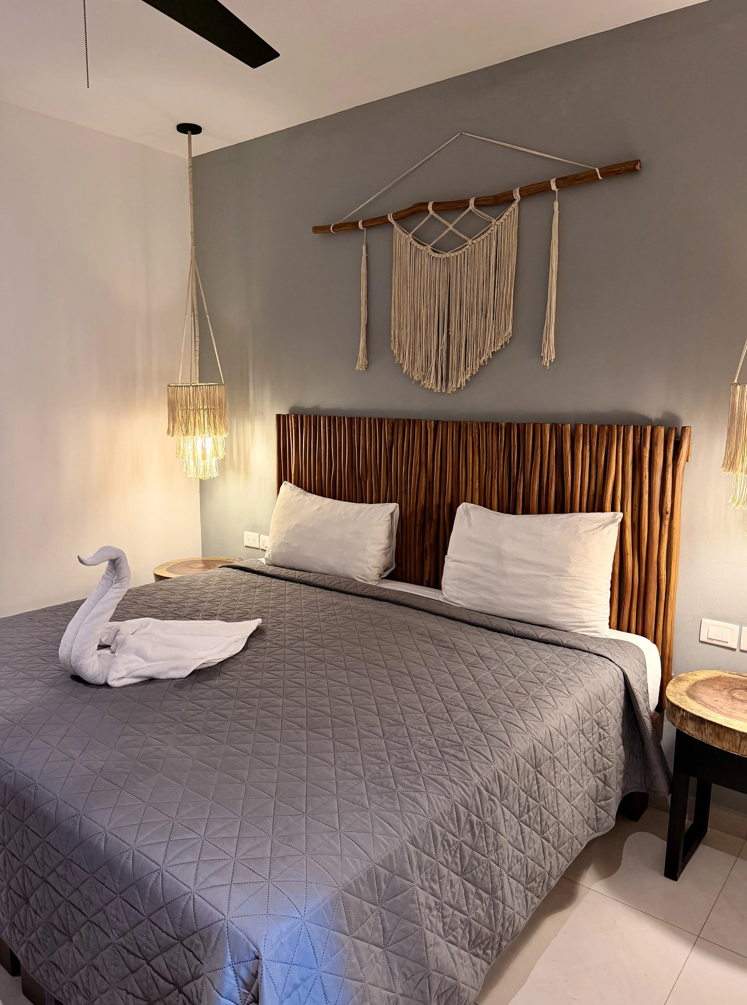 Cama con dos almohadas blancas, cubrecama gris con diseño de rombos, y una toalla doblada en forma de cisne en la cama, en un dormitorio con paredes blancas y una pared gris, con un cabecero de madera y cortinas de lona color beige. Encima, en la par